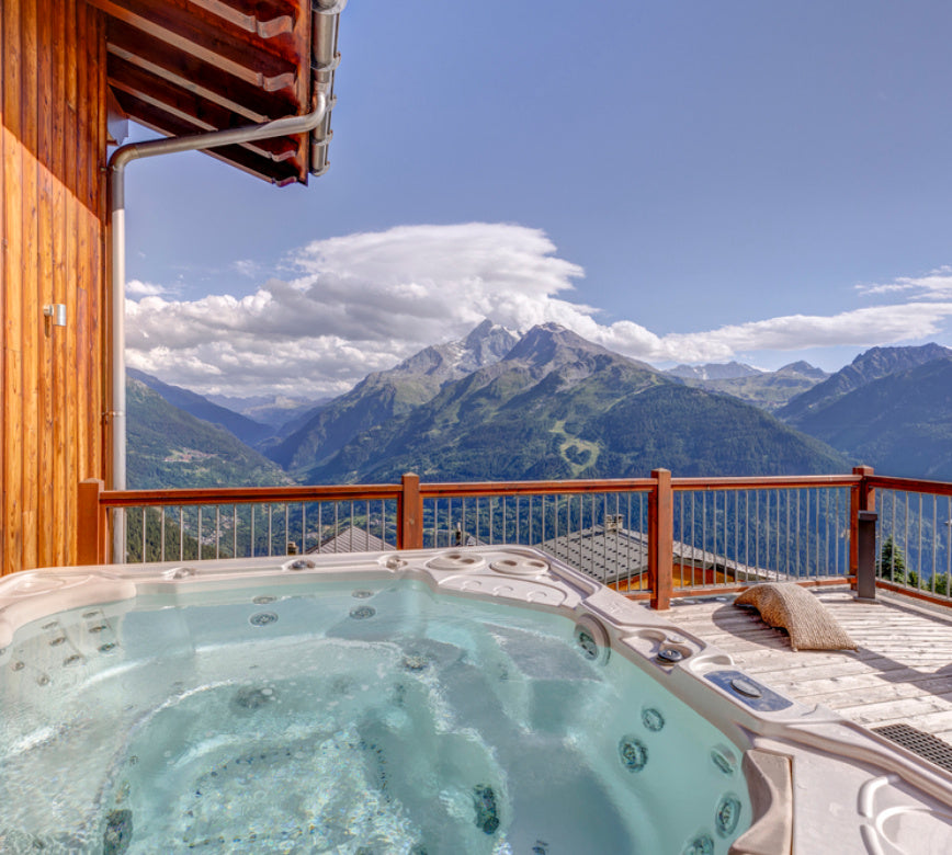 Un jacuzzi extérieur rempli d’eau claire occupe le premier plan, installé sur une terrasse en bois avec un garde-corps.
Au second plan, de grandes montagnes verdoyantes s’élèvent sous un ciel bleu lumineux, avec des nuages blancs qui entourent les sommets.
