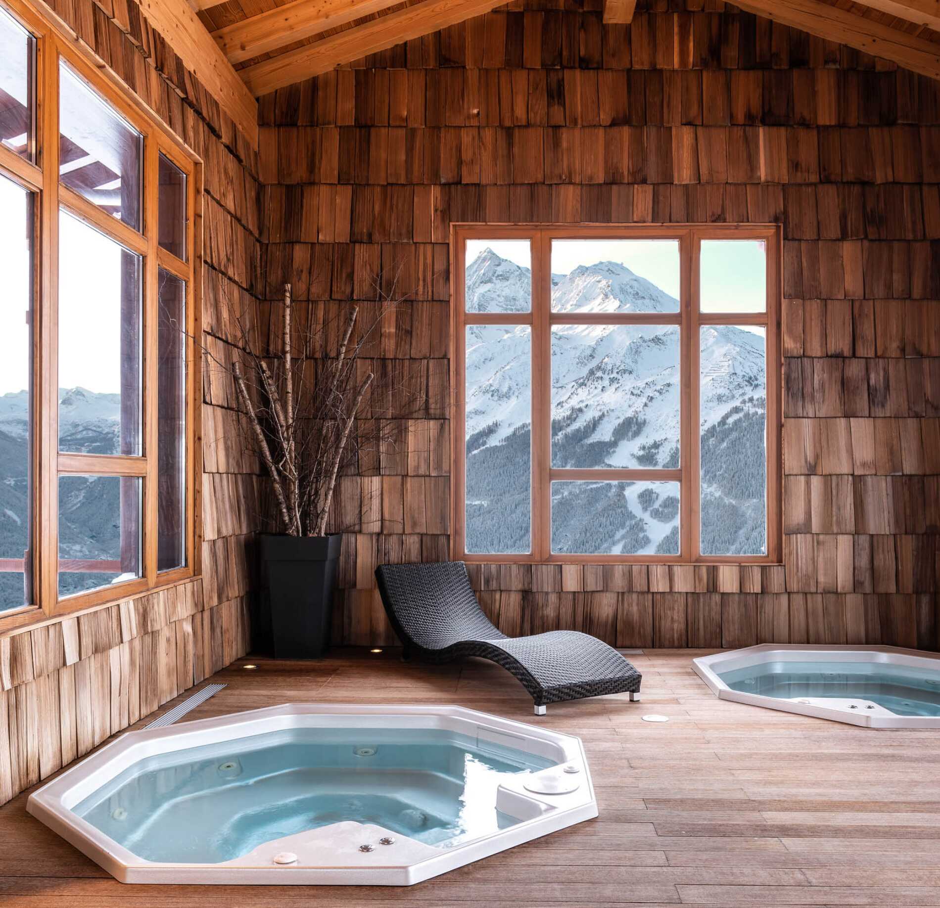 Une pièce intérieure entièrement en bois abrite deux petits jacuzzis encastrés dans le sol, avec une chaise longue tressée posée entre les deux.
De grandes fenêtres laissent apparaître des montagnes enneigées à l’extérieur, apportant une lumière naturelle qui éclaire le bois clair et les surfaces.