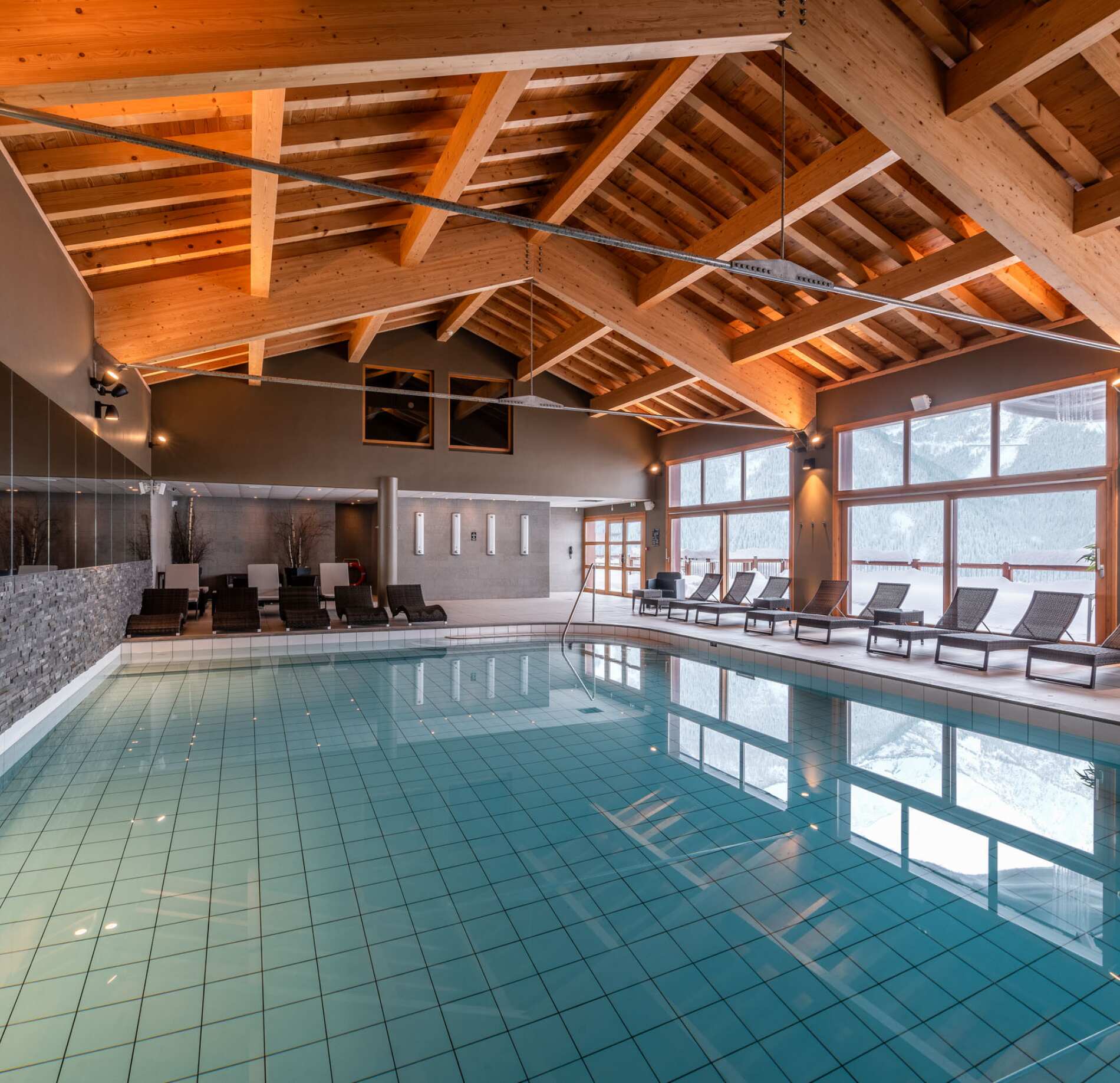 Dans le spa de l'I.L.Y hotels la roisiere , il y a une grande piscine intérieure s’étend au centre d’une pièce spacieuse avec un sol carrelé et une charpente en bois apparente au plafond.
De larges fenêtres laissent entrer la lumière naturelle et donnent sur un paysage enneigé, tandis que des transats sont alignés le long des baies vitrées.