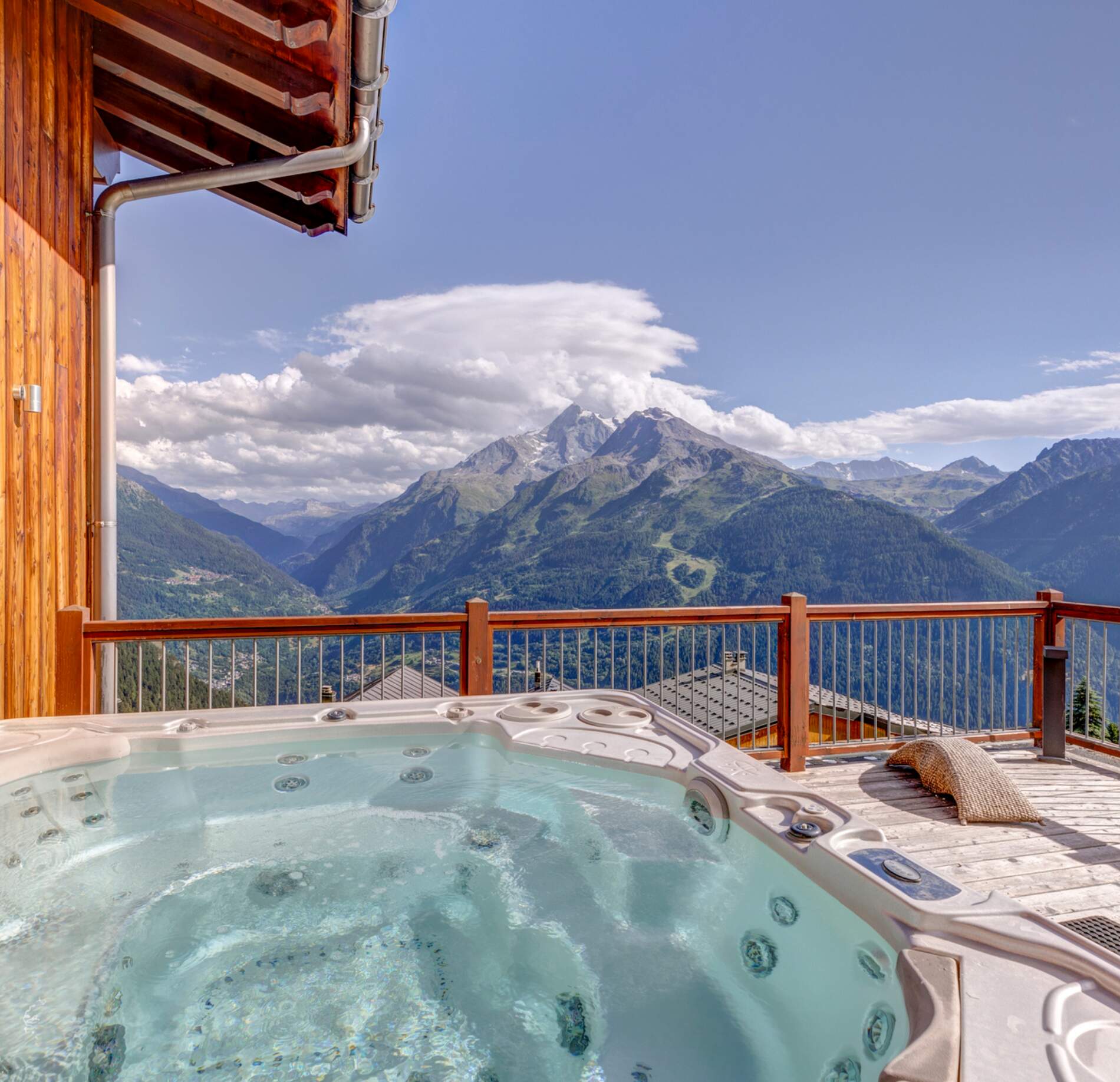 Il y a un jacuzzi extérieur qui occupe le premier plan, installé sur une terrasse en bois avec un garde-corps.
Au second plan, de grandes montagnes verdoyantes s’élèvent sous un ciel bleu lumineux, avec des nuages blancs qui entourent les sommets.