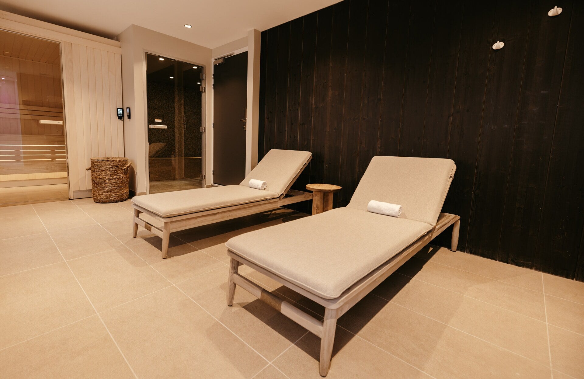 Un espace de détente intime et élégant, avec deux chaises longues en bois clair surmontées de coussins épais et de serviettes soigneusement roulées.
À l’arrière, un sauna lumineux et une cabine vitrée créent un parcours bien‑être complet, dans une atmosphère chaleureuse et parfaitement aménagée pour le repos.