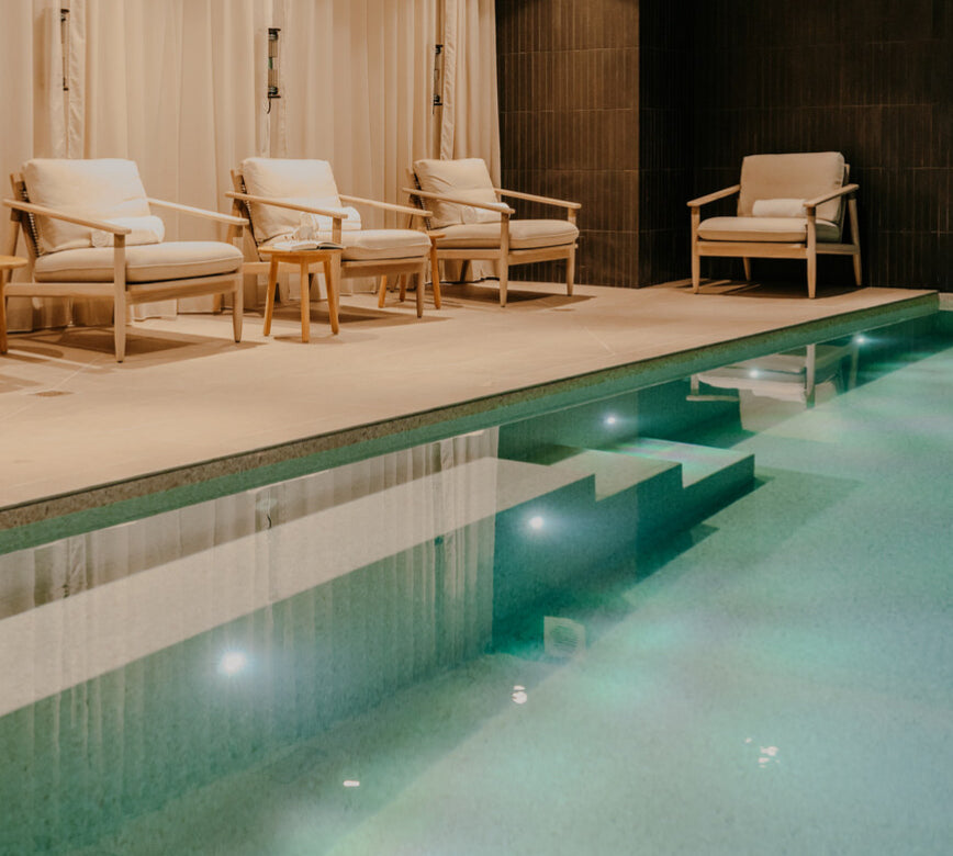 Une piscine intérieure du spa de la maison le Goyen s’étend au premier plan, avec une eau claire éclairée par des lumières intégrées et des marches visibles sous la surface.
Le long du bassin, plusieurs fauteuils confortables en bois clair sont alignés contre un rideau beige, créant un espace de repos calme et élégant.