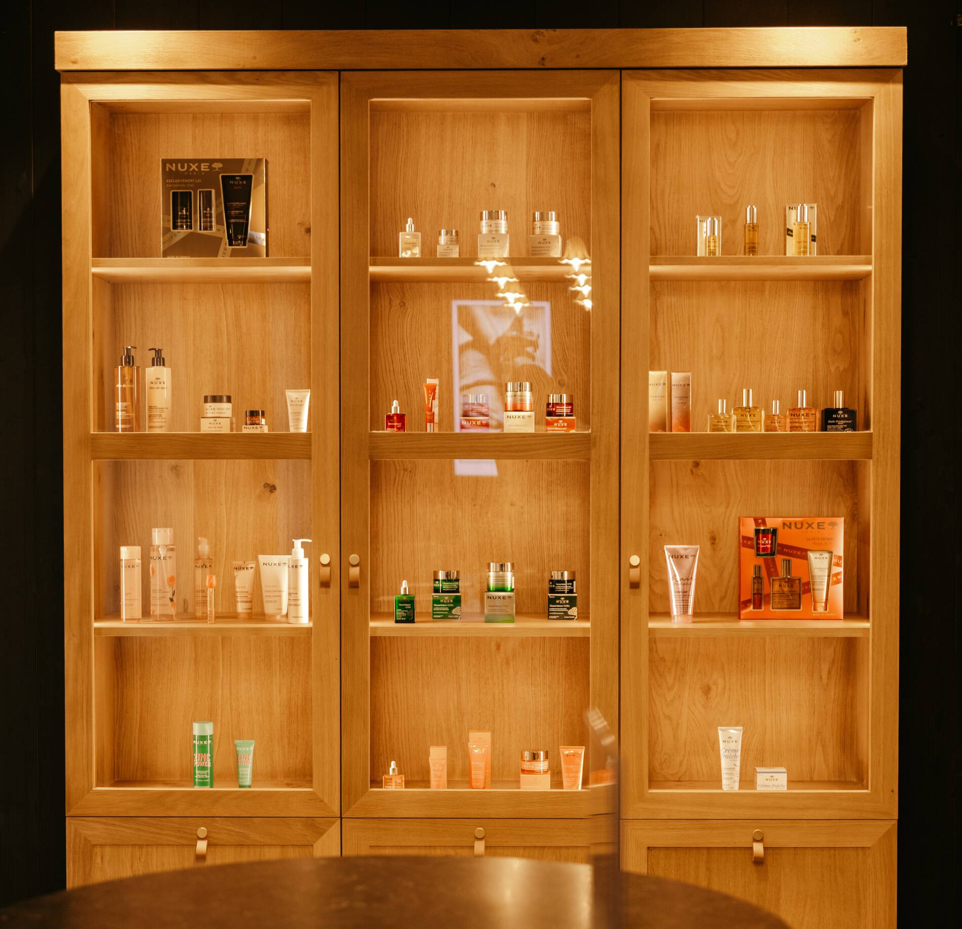 Une vitrine en bois du spa de la maison le Goyen présente plusieurs produits de cosmétique NUXE soigneusement rangés sur des étagères éclairées.
Les flacons et tubes, aux couleurs variées, sont disposés de façon ordonnée derrière des portes vitrées, créant une ambiance chaleureuse et soignée.