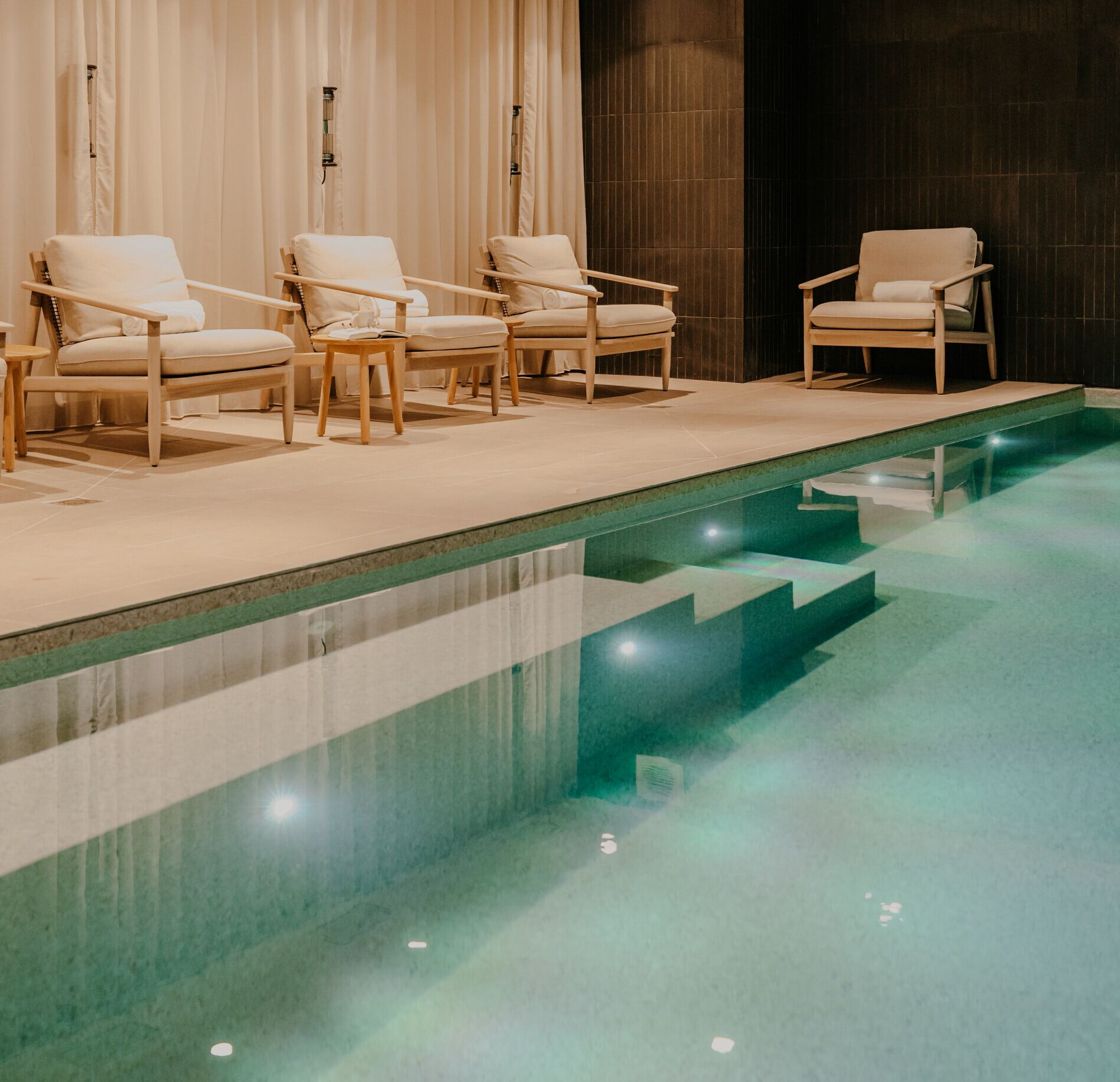 Une piscine intérieure du spa de la maison le Goyen s’étend au premier plan, avec une eau claire éclairée par des lumières intégrées et des marches visibles sous la surface.
Le long du bassin, plusieurs fauteuils confortables en bois clair sont alignés contre un rideau beige, créant un espace de repos apaisant et élégant.