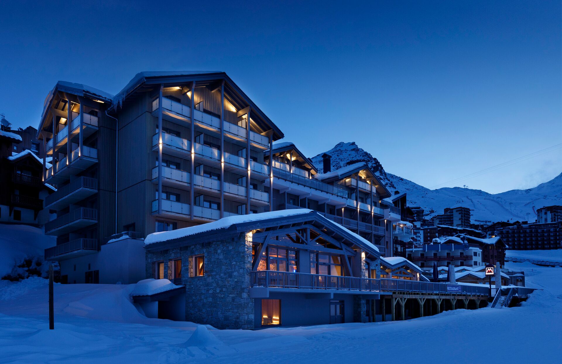 Un élégant hôtel de montagne nommé Le Marielle, baigné dans la lumière bleutée du crépuscule, se distingue par son architecture contemporaine mêlant bois et pierre. Entouré de sommets enneigés, l’établissement dégage une atmosphère chaleureuse grâce à ses lumières tamisées, offrant un cadre idéal pour une escapade luxueuse au cœur des Alpes.