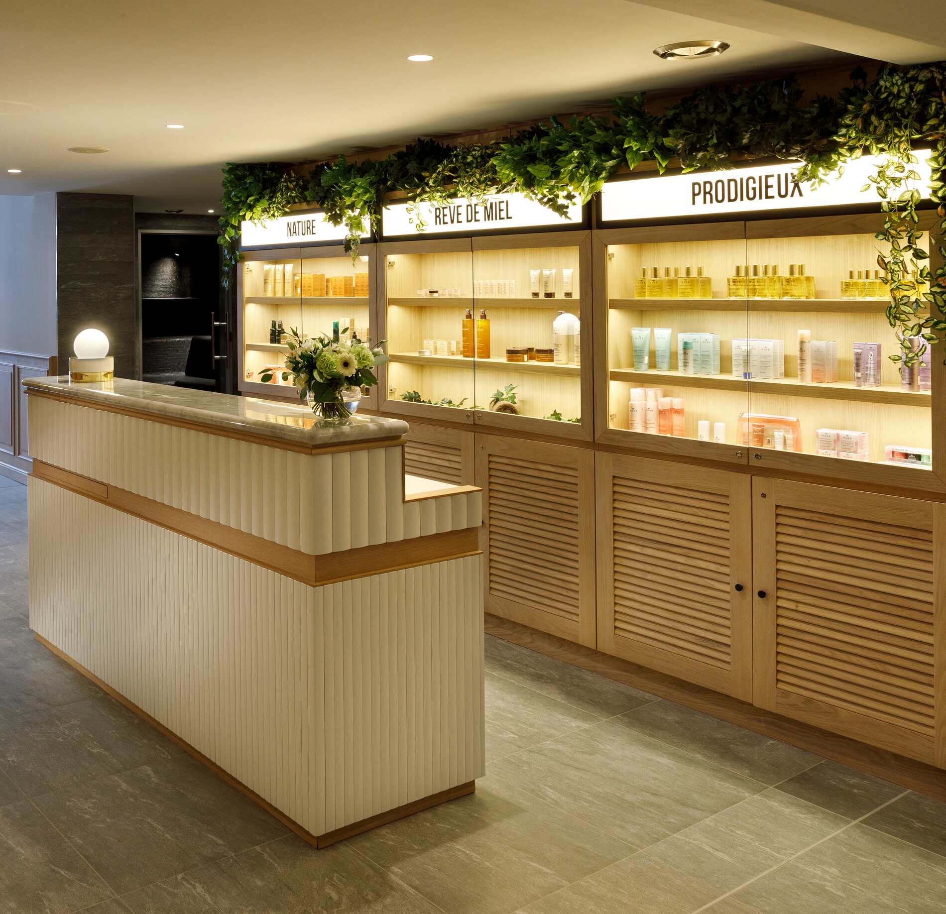 Un espace d’accueil du spa de l'hotel Marielle présente un comptoir en bois clair face à des étagères éclairées remplies de produits de soin bien alignés.
Au-dessus, des panneaux indiquent « NATURE », « RÊVE DE MIEL » et « PRODIGIEUX », avec des plantes vertes décoratives créant une atmosphère naturelle et élégante.