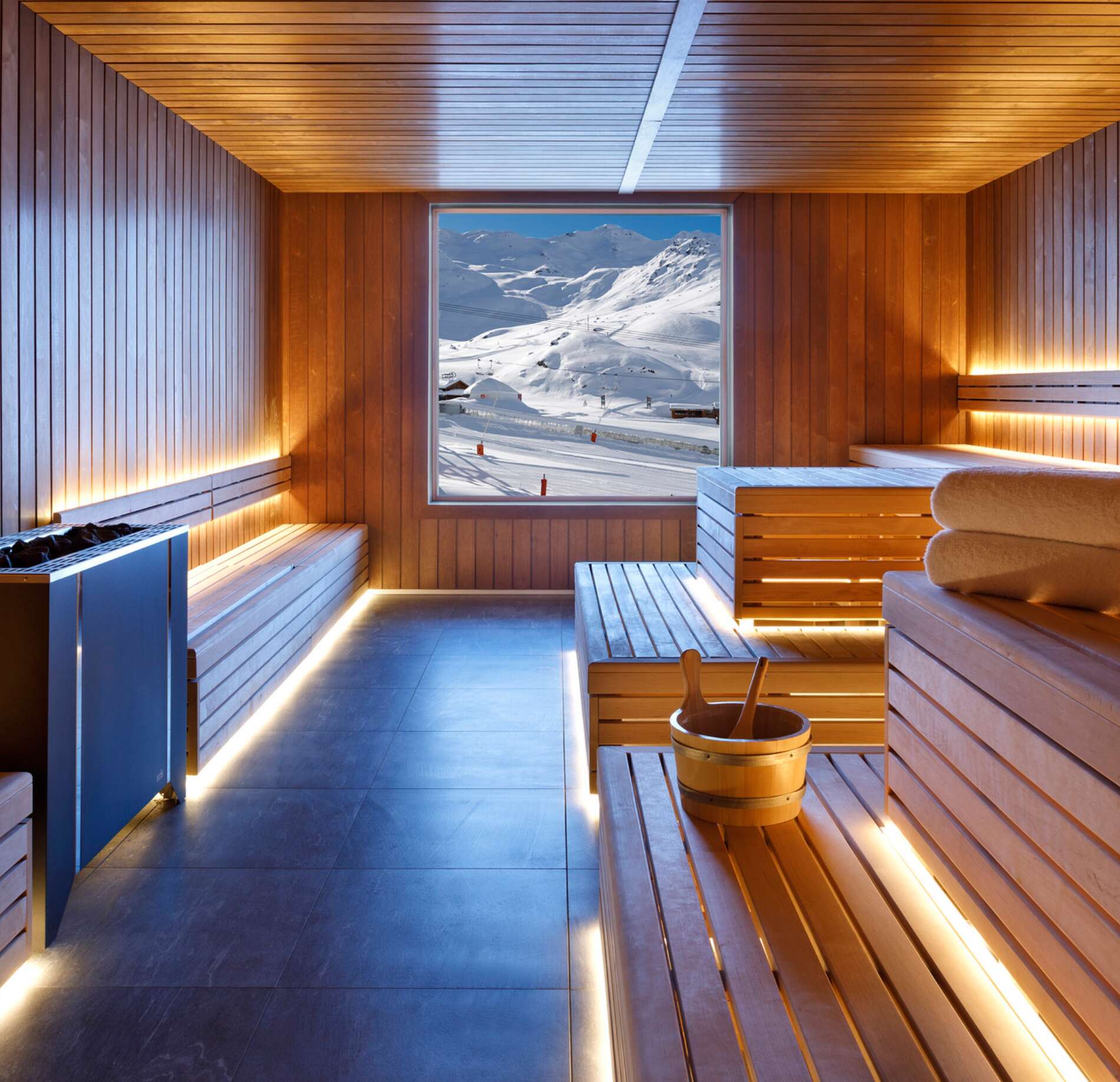 Un sauna du spa de l'hotel Marielle entièrement habillé de bois clair dispose de bancs éclairés par une lumière douce intégrée.
Au centre, une grande fenêtre encadre une vue sur des montagnes enneigées, tandis qu’un seau en bois avec louche est posé sur le banc, renforçant l’ambiance chaleureuse et apaisante.