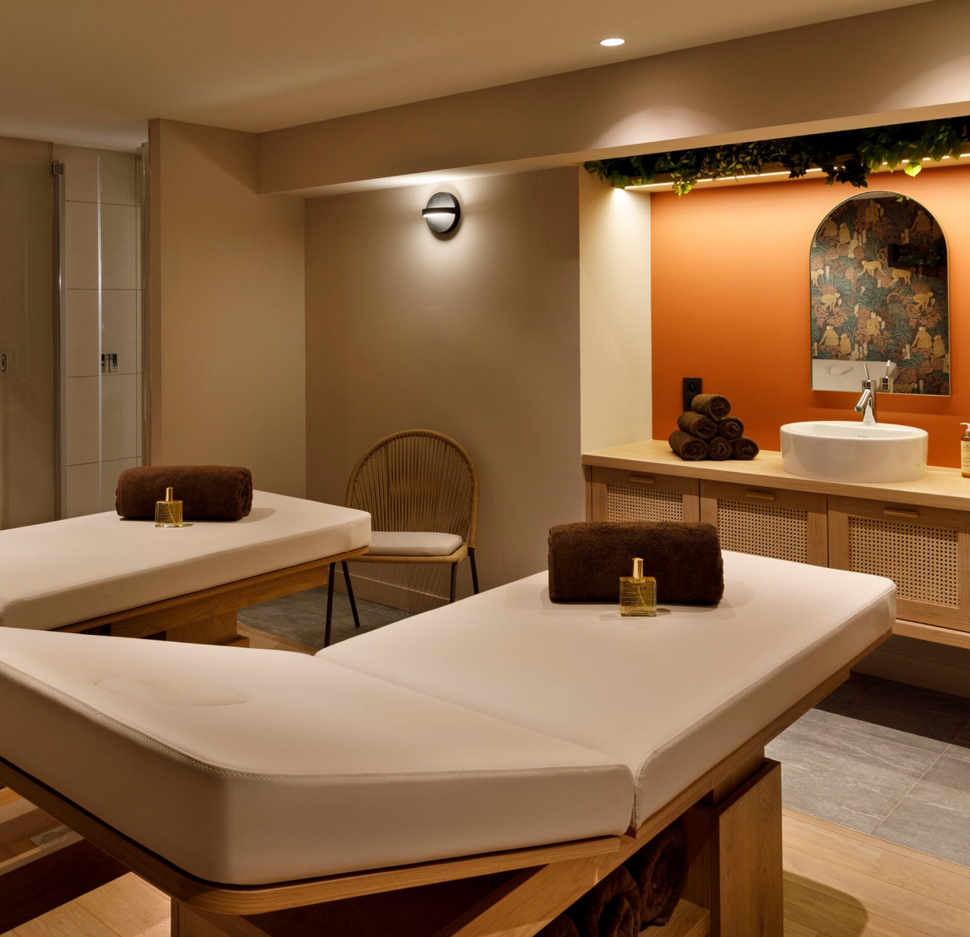 Une salle de soins du spa de l'hotel Marielle présente deux tables de massage recouvertes de draps blancs, avec des serviettes roulées et des huiles posées dessus.
À l’arrière, un meuble en bois avec une vasque, des serviettes empilées et un mur orangé décoratif créent une ambiance chaleureuse et soignée.
