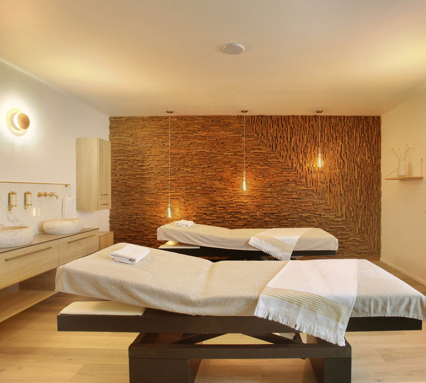 Une salle de soins du spa le Negrecoste présente deux tables de massage recouvertes de linges clairs, avec des serviettes soigneusement disposées au centre.  
Au fond, un mur en bois texturé avec suspensions lumineuses crée une ambiance douce et chaleureuse, complétée par un meuble avec double vasque sur le côté.