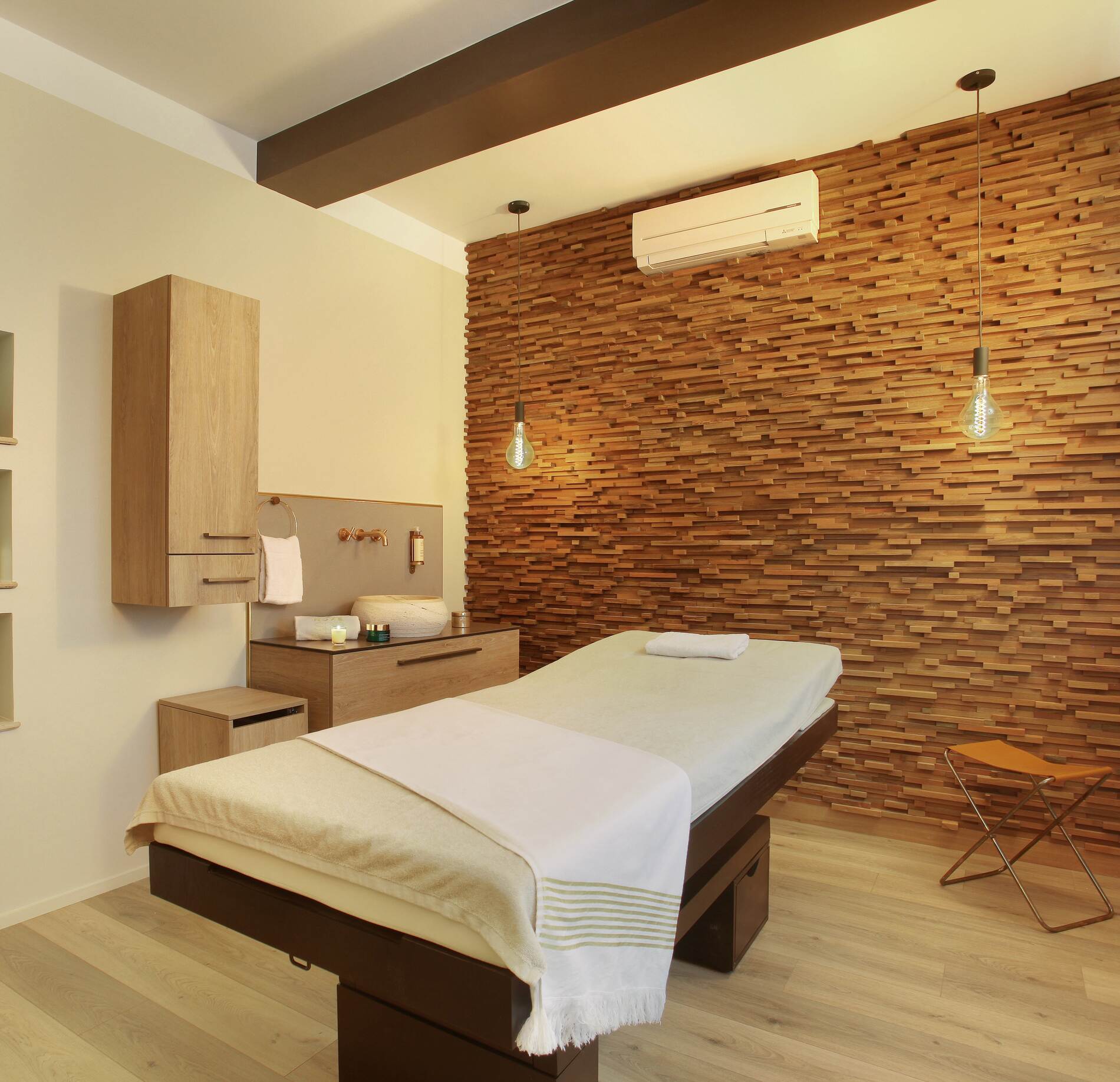 Une cabine de soins du spa le Negrecoste présente une table de massage unique recouverte de linges clairs, avec une serviette soigneusement pliée au centre.
Le mur en bois texturé, les suspensions lumineuses et le mobilier en bois clair créent une ambiance chaleureuse et épurée, complétée par un petit espace avec vasque sur le côté.