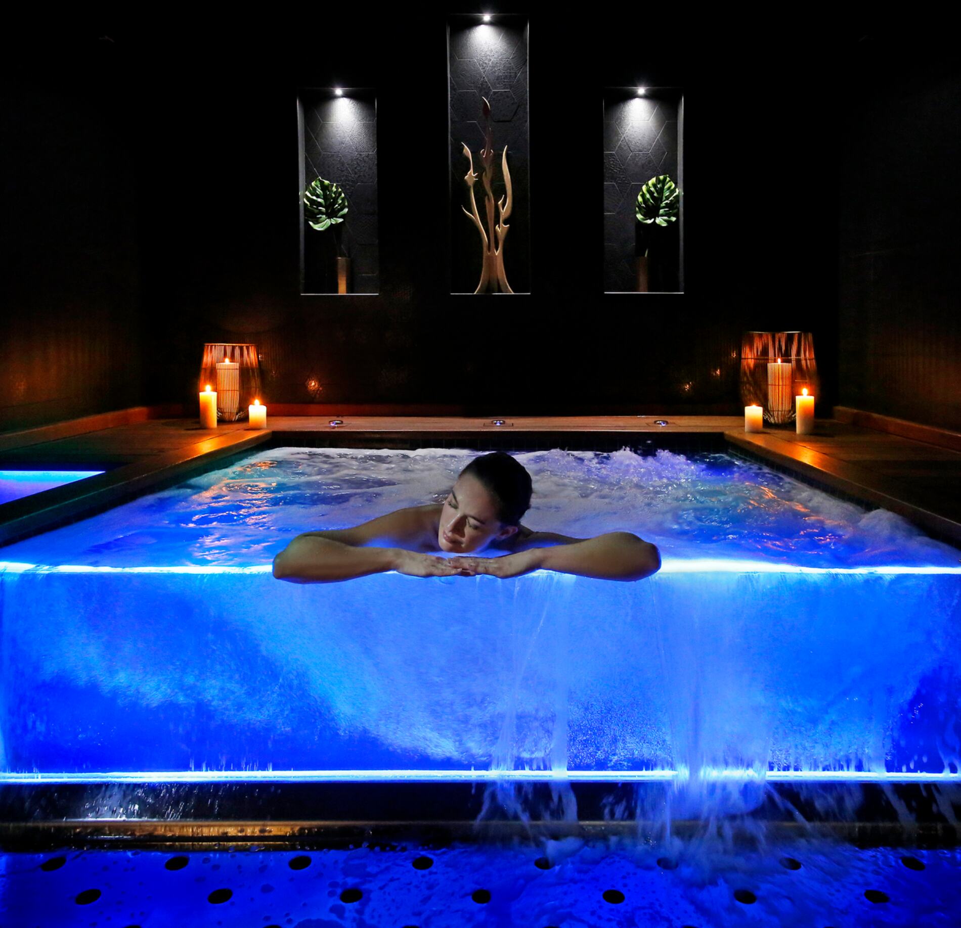 Un bassin à remous du spa le Negrecoste est éclairé d’une lumière bleue, avec de l’eau en mouvement et une personne appuyée sur le bord au centre de l’image.
Autour, des bougies et des niches murales décorées de plantes et d’objets créent une ambiance sombre, intime et relaxante.