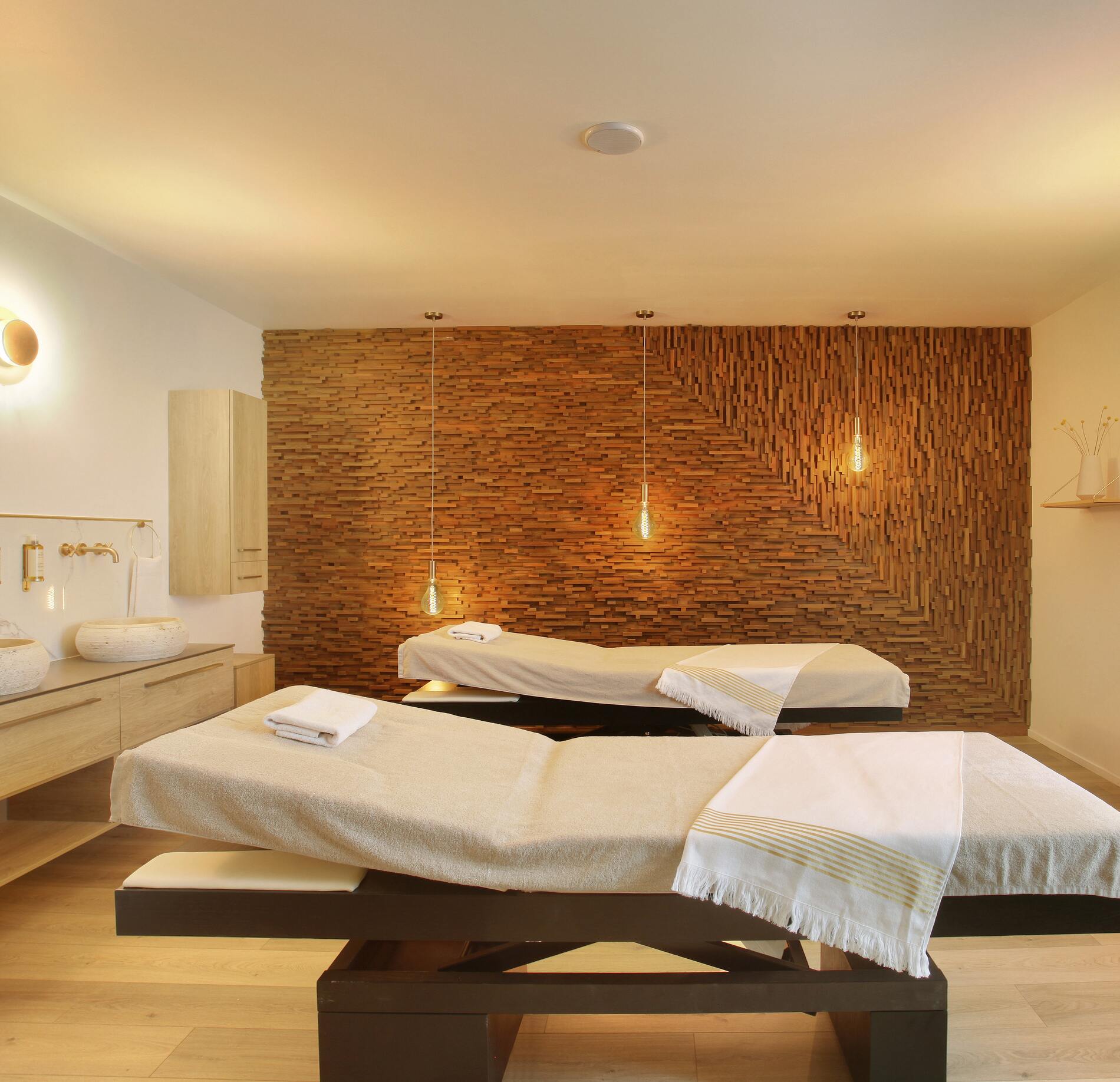 Une salle de soins du spa le Negrecoste présente deux tables de massage recouvertes de linges clairs, avec des serviettes soigneusement disposées au centre.  
Au fond, un mur en bois texturé avec suspensions lumineuses crée une ambiance douce et chaleureuse, complétée par un meuble avec double vasque sur le côté.