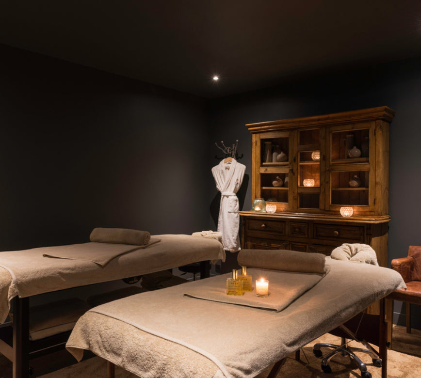 Une cabine de soins du spa les pins de Cesar présente deux tables de massage recouvertes de linges clairs avec des serviettes roulées posées dessus.
À l’arrière, un meuble en bois décoratif avec bougies, un peignoir suspendu et un éclairage tamisé créent une ambiance chaleureuse et intimiste.