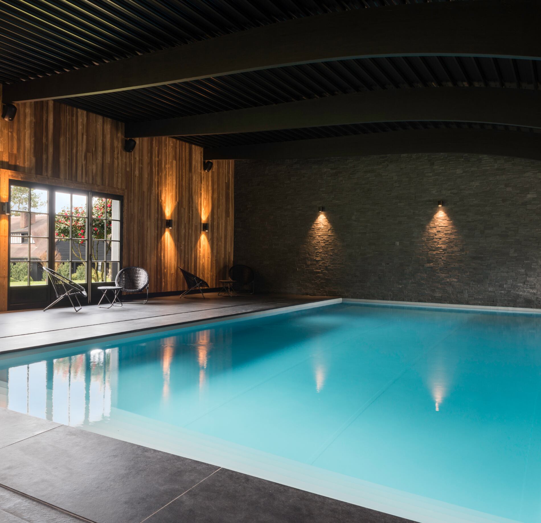 Une piscine intérieure du spa les pins de Cesar s’étend dans une grande pièce aux murs en bois et en pierre, avec une eau calme aux reflets bleu clair.
Des fauteuils sont disposés près d’une porte vitrée donnant sur l’extérieur, tandis que des éclairages muraux créent une ambiance douce et relaxante.
