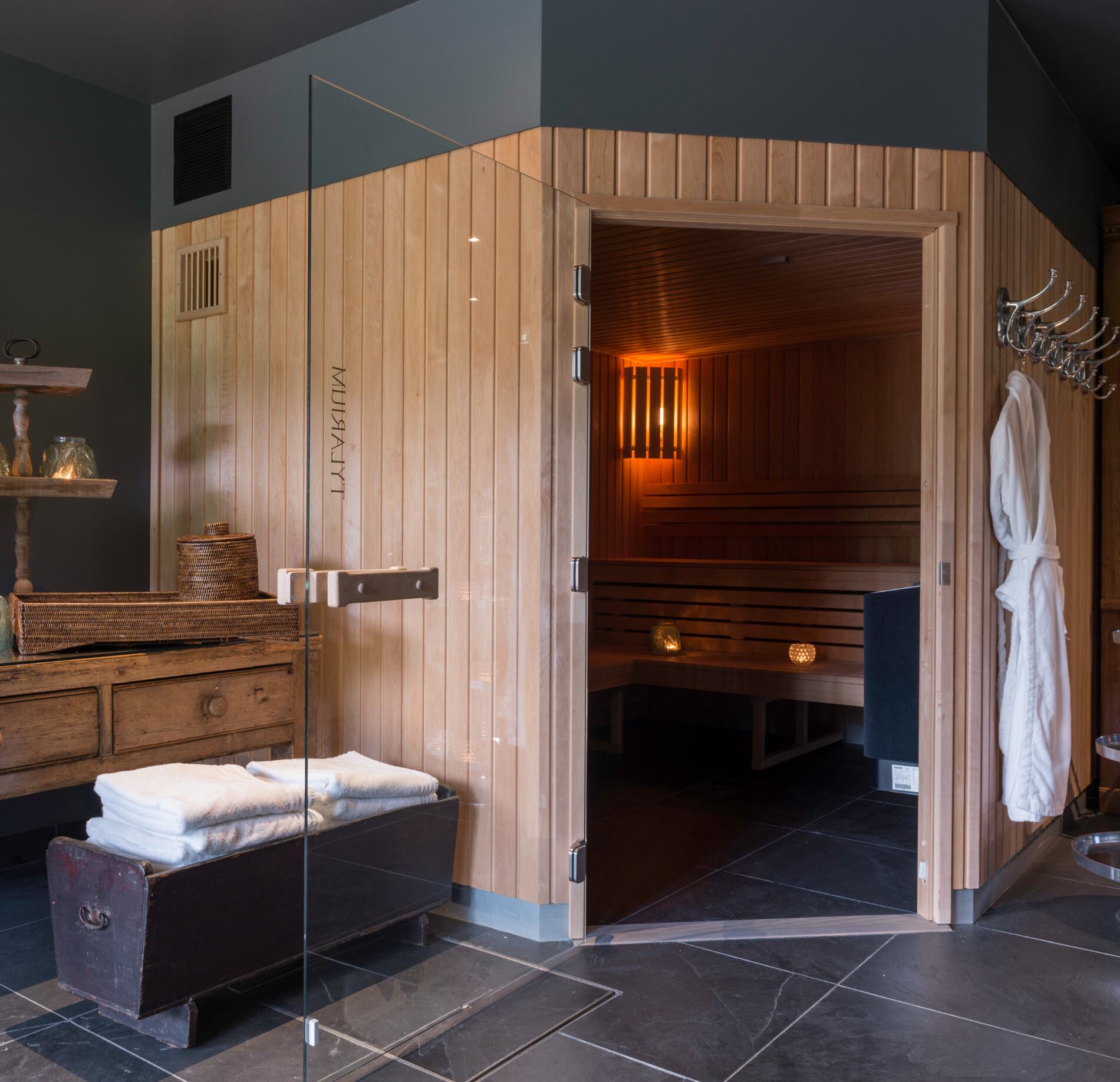 Un sauna du spa les pins de Cesar en bois clair est ouvert, laissant apparaître des bancs et un éclairage chaud à l’intérieur.
À l’extérieur, des serviettes pliées, un meuble en bois et un peignoir suspendu complètent cet espace avec une ambiance chaleureuse et authentique.