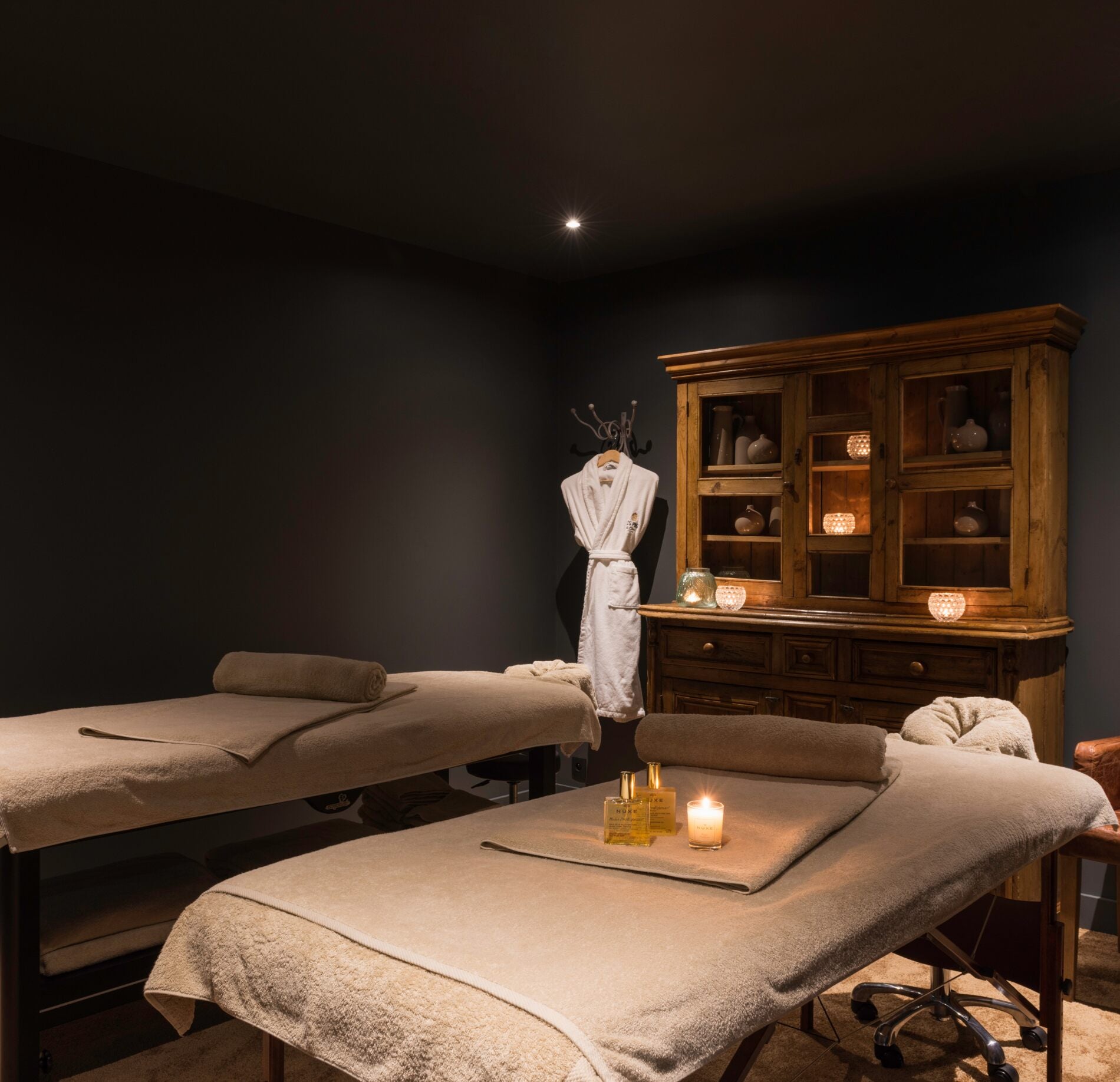 Une cabine de soins du spa les pins de Cesar présente deux tables de massage recouvertes de linges clairs avec des serviettes roulées posées dessus.
À l’arrière, un meuble en bois avec bougies, un peignoir suspendu et un éclairage tamisé créent une ambiance chaleureuse et intimiste.