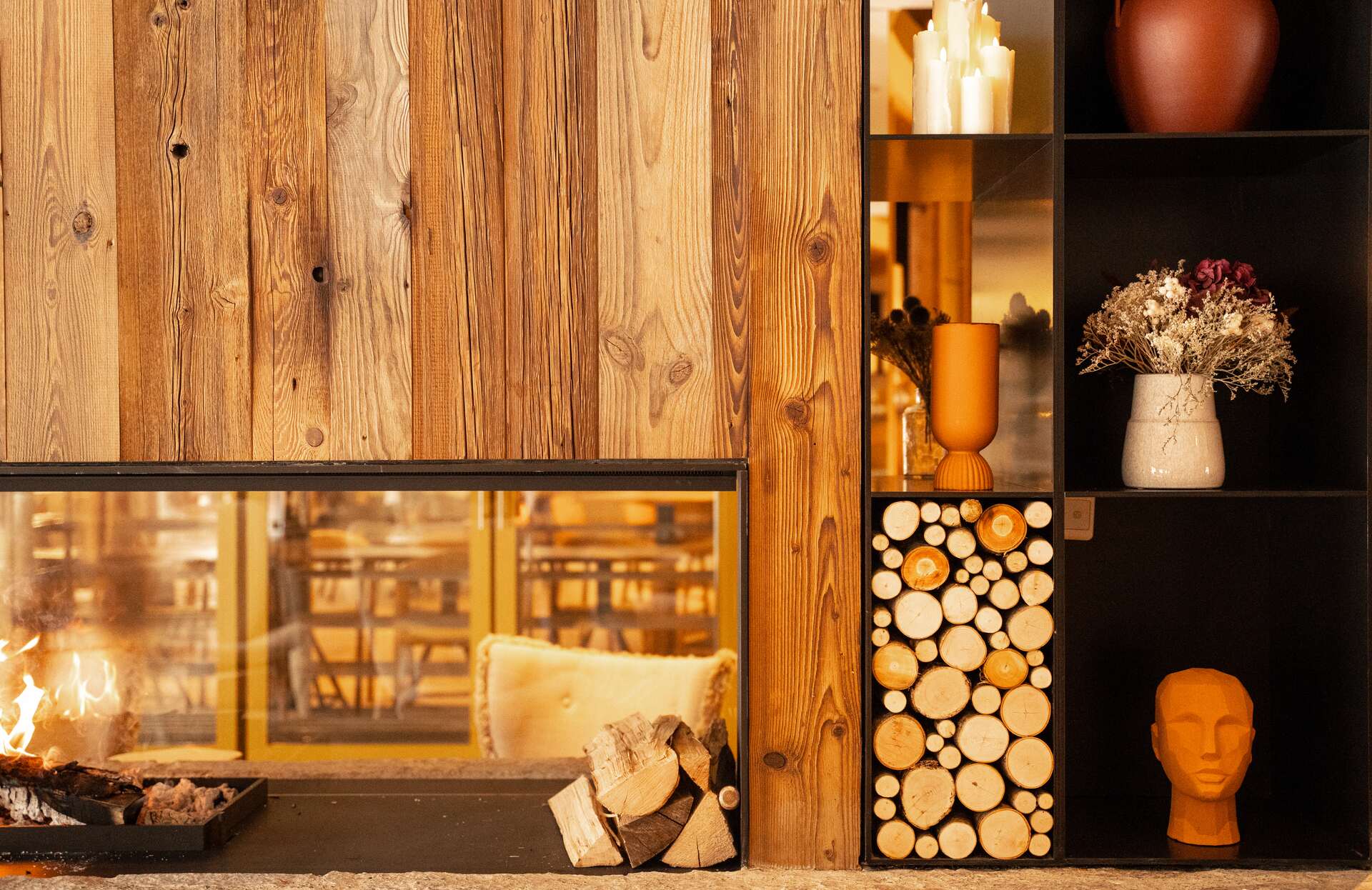 Un espace chaleureux dominé par une cheminée encastrée met en valeur des flammes douces derrière une vitre, entourée de bois brut aux nuances naturelles.
À droite, des étagères sombres décorées de bougies, de vases et de rondins soigneusement disposés créent une ambiance élégante et enveloppante, invitant à une détente absolue.