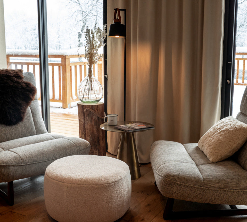 Un coin détente chaleureux de l'Hotel Spa Loudenvielle, avec deux fauteuils confortables en tissu clair disposés autour d’une petite table ronde.
Face à une grande baie vitrée ouverte sur un balcon enneigé, une lampe douce et des rideaux épais créent une atmosphère cosy et apaisante.