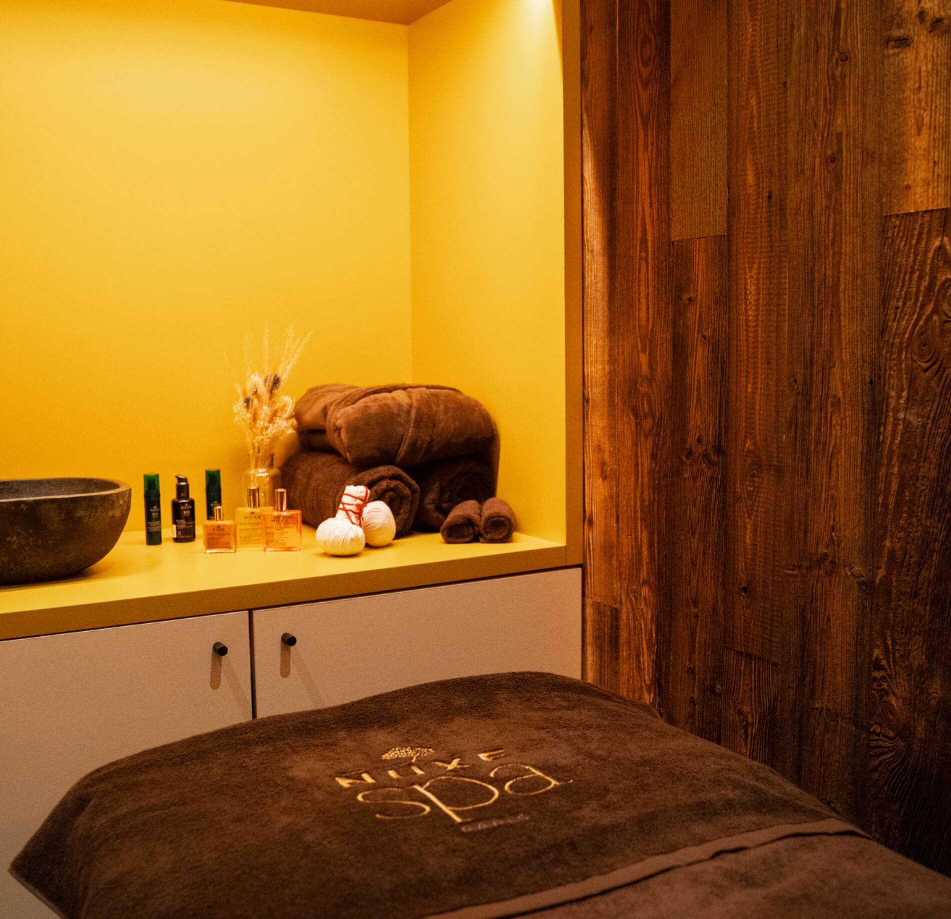 Une cabine de soin chaleureuse de l'Hotel Spa Loudenvielle, avec une table de massage recouverte d’un linge brun et entourée de produits de soin disposés avec précision.
Le contraste entre le mur jaune lumineux et le bois foncé crée une ambiance enveloppante, tandis que les serviettes pliées renforcent l’atmosphère cosy et soignée.