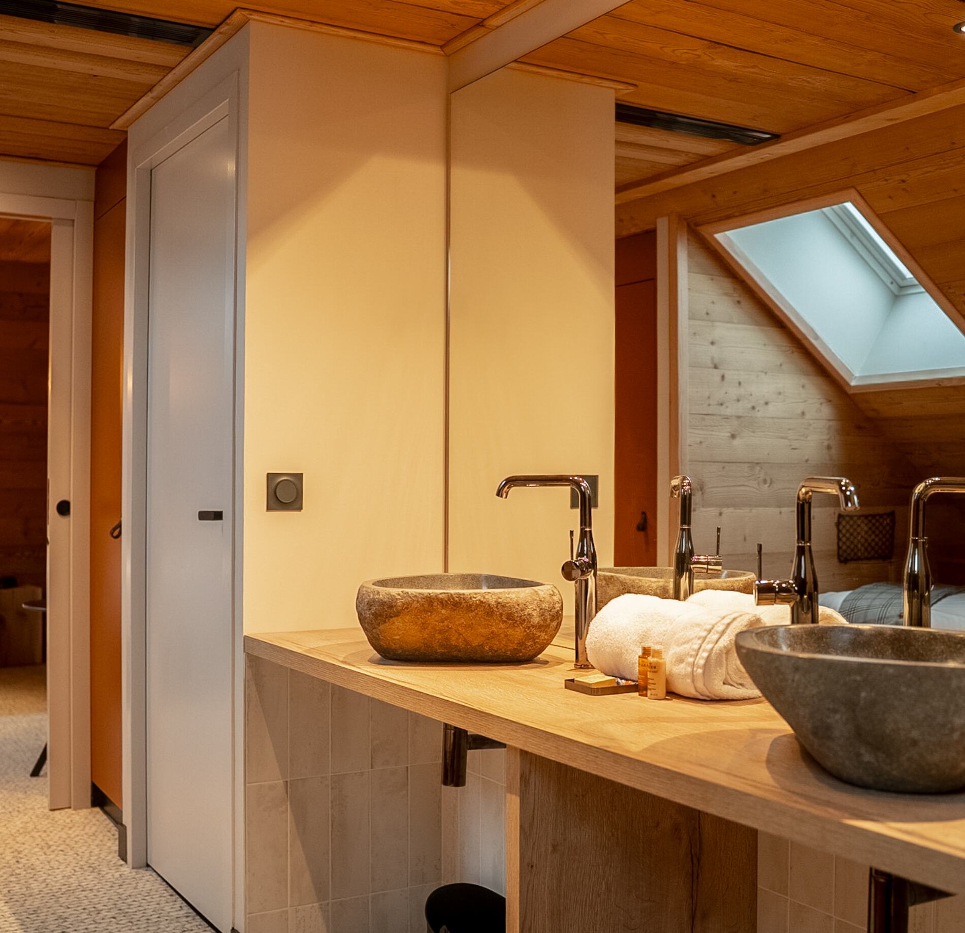 Une salle de bain moderne de l'Hotel Spa Loudenvielle, avec un plan de toilette en bois surmonté de deux vasques en pierre et de robinets métalliques élégants.
Le décor mêle murs clairs et bois chaleureux, avec un grand miroir et une fenêtre de toit laissant entrer une lumière douce dans une atmosphère apaisante.