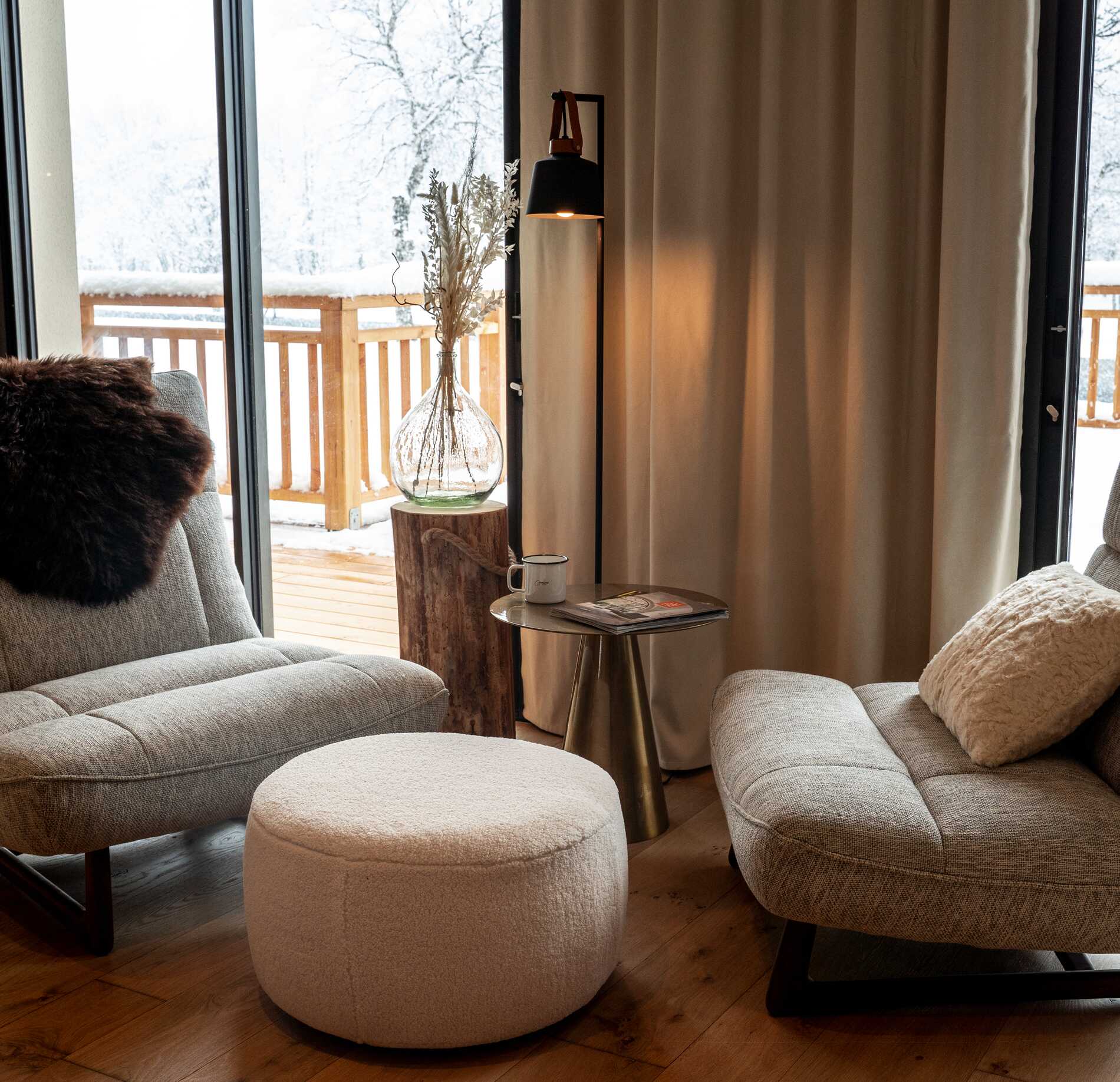 Un coin détente chaleureux de l'Hotel Spa Loudenvielle, avec deux fauteuils confortables en tissu clair disposés autour d’une petite table ronde.
Face à une grande baie vitrée donnant sur un balcon enneigé, une lampe douce et des rideaux épais créent une atmosphère cocooning et apaisante.