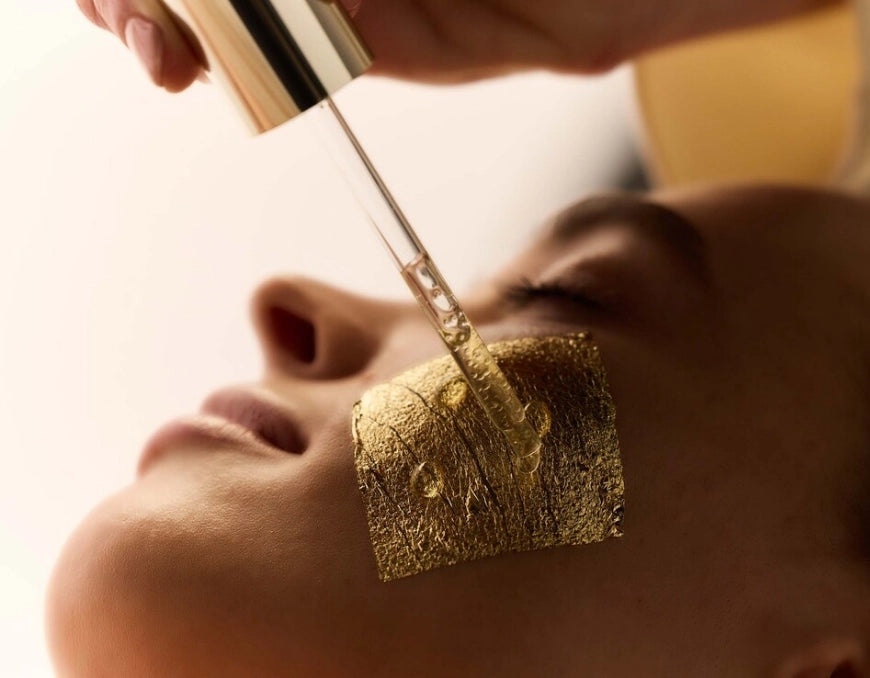 Femme allongée, yeux fermés, une feuille d'or sur la pommette sur laquelle une praticienne dépose quelques gouttes de sérum doré à l'aide d'une pipette
