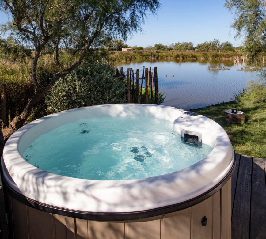 Un jacuzzi extérieur du spa de l'hotel Mas de la Fouque est installé sur une terrasse en bois, rempli d’une eau claire légèrement agitée.
Face à lui, un étang naturel entouré de végétation et d’arbres crée un cadre sauvage et paisible sous un ciel dégagé.