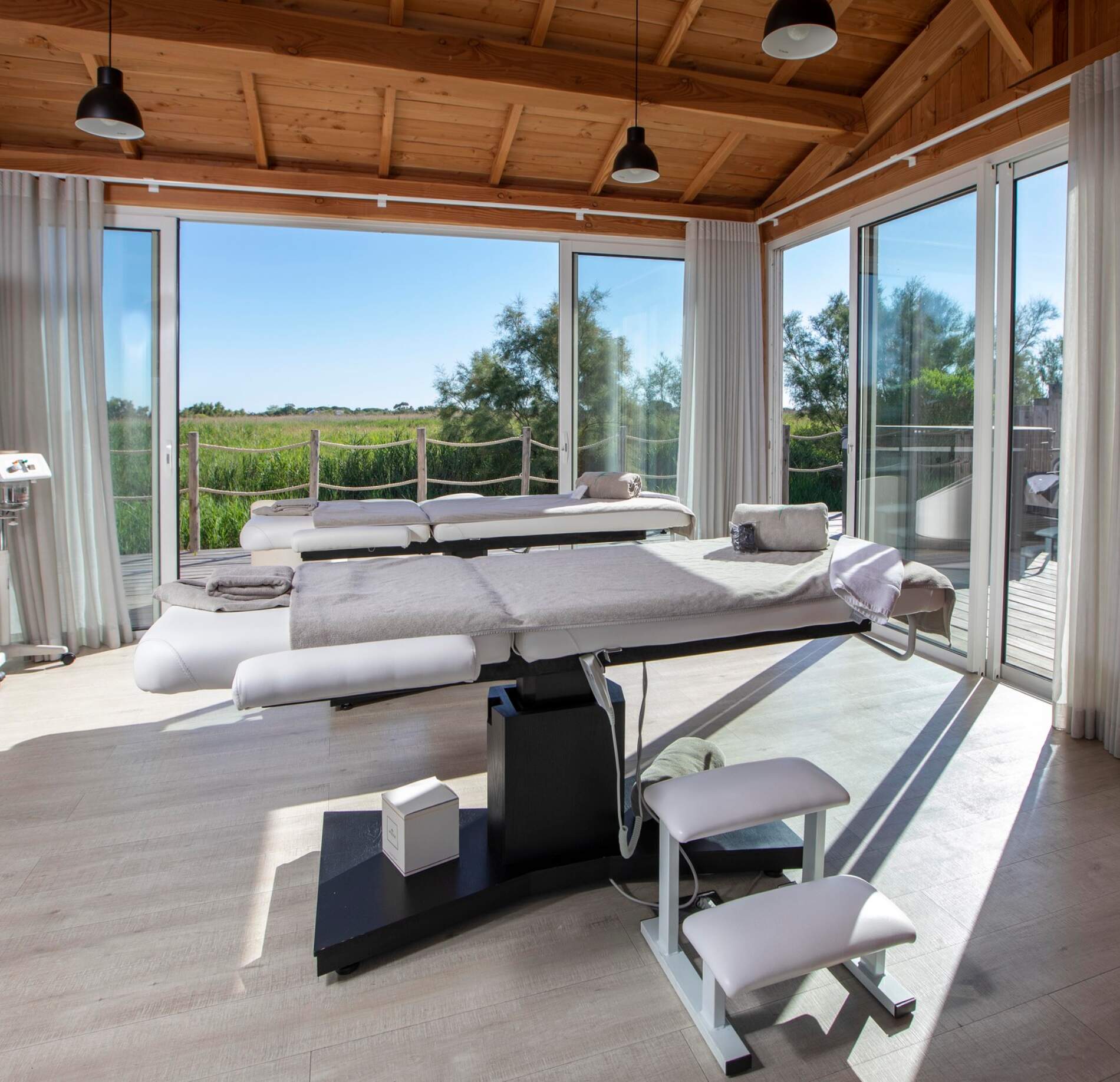 Une cabine de soins lumineuse du spa de l'hotel Mas de la Fouque présente deux tables de massage recouvertes de linges clairs, avec serviettes roulées et coussins disposés dessus.
De grandes baies vitrées entourent la pièce et offrent une vue dégagée sur la nature et la végétation, baignant l’espace de lumière naturelle.
