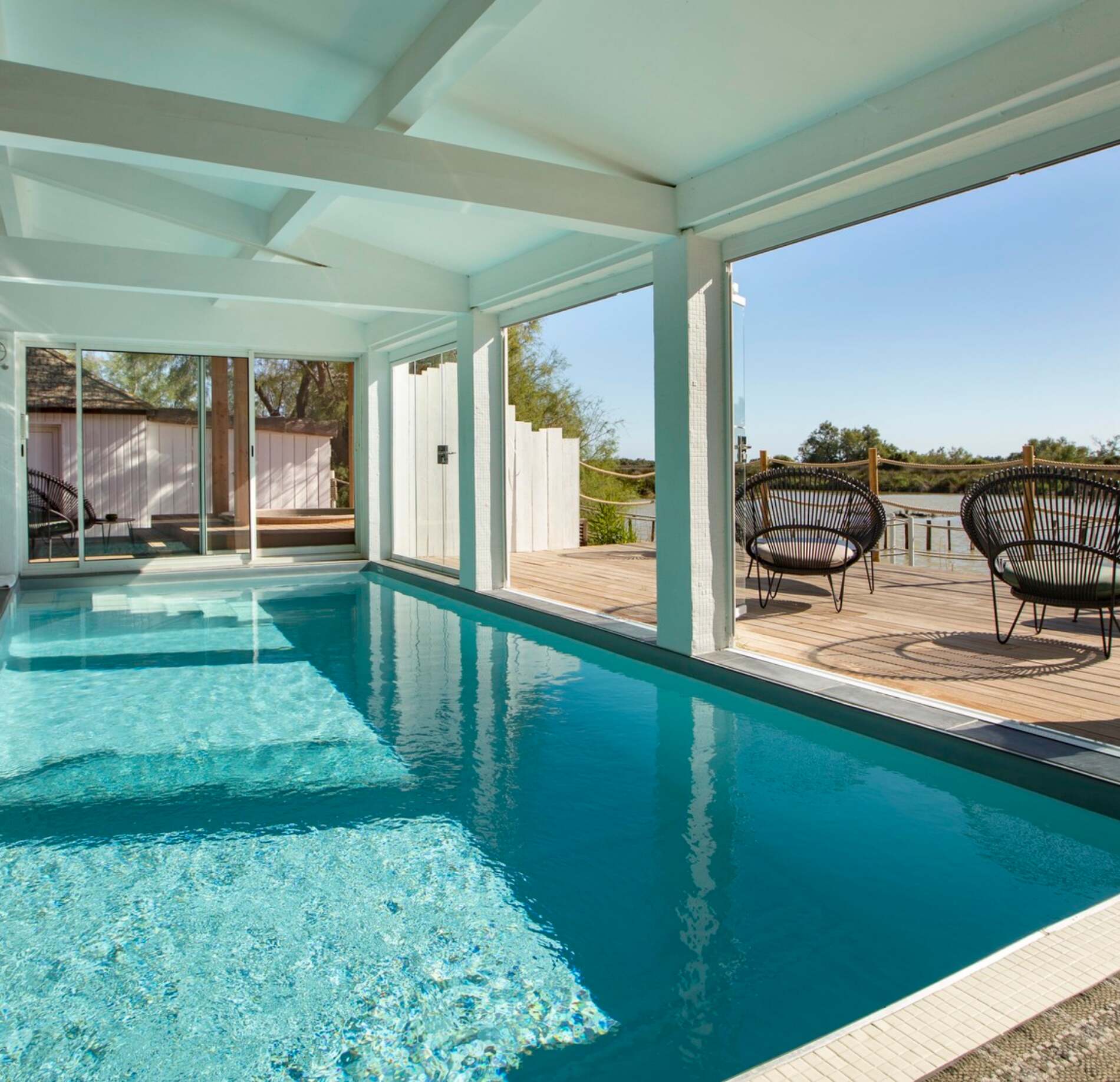 Une piscine intérieure du spa de l'hotel Mas de la Fouque s’étend sous une structure blanche, avec une eau turquoise limpide occupant tout le premier plan.
De grandes ouvertures donnent sur une terrasse en bois avec des fauteuils ronds et une vue sur la nature, baignant l’espace de lumière naturelle et apaisante.