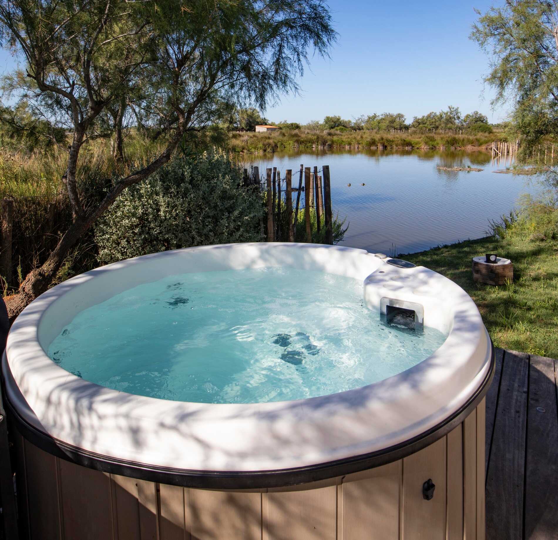 Un jacuzzi extérieur du spa de l'hotel Mas de la Fouque est installé sur une terrasse en bois, rempli d’une eau claire légèrement agitée.
Face à lui, un étang naturel entouré de végétation et d’arbres crée un cadre sauvage et paisible sous un ciel dégagé.