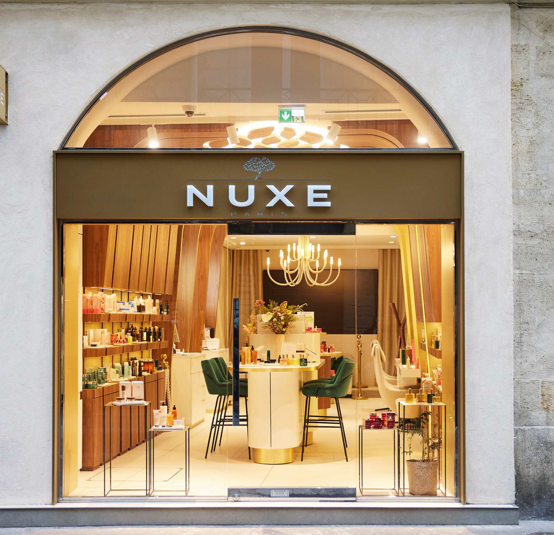 Une vitrine de boutique élégante est encadrée par une grande arche en verre, avec le nom « NUXE » affiché en lettres blanches au-dessus de l’entrée. L’intérieur est très lumineux, laissant apparaître un espace organisé et chaleureux.
À l’intérieur, des étagères en bois présentent de nombreux produits, tandis qu’un comptoir central entouré de chaises vertes attire le regard. Un lustre suspendu diffuse une lumière douce, renforçant l’ambiance raffinée et accueillante de la boutique.