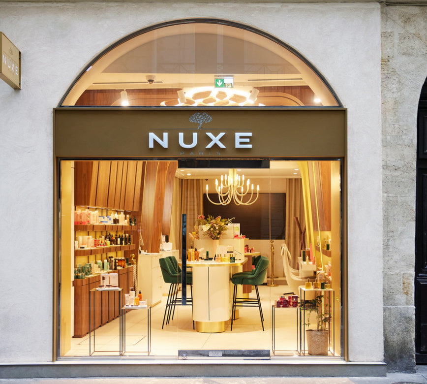 Une vitrine élégante donne sur une boutique lumineuse, avec une grande arche en verre encadrant l’entrée. Le mot « NUXE » est inscrit en lettres blanches au-dessus de la porte, bien visible depuis l’extérieur.
À l’intérieur, un espace chaleureux est aménagé avec des étagères en bois remplies de produits, un comptoir central et des chaises hautes vertes. Un lustre suspendu éclaire la pièce, créant une ambiance raffinée et accueillante.