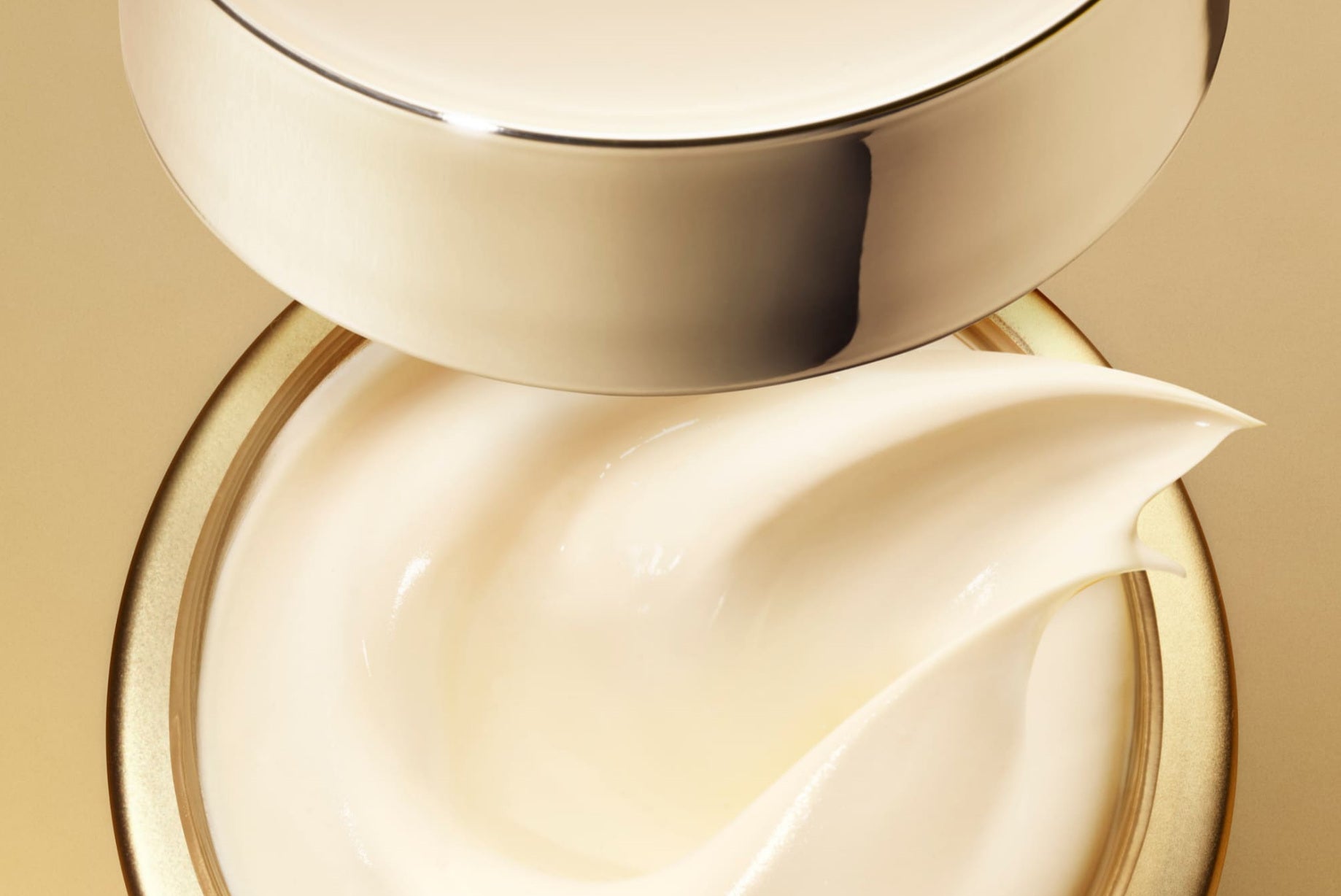 Un pot de crème ouvert est vu de très près, avec un couvercle métallique doré suspendu juste au-dessus. À l’intérieur, la crème blanche forme un mouvement fluide et sculpté, avec une pointe relevée qui attire le regard.
La matière apparaît lisse, épaisse et brillante, reflétant la lumière chaude autour d’elle. Le fond doré renforce l’aspect luxueux et met en valeur la texture soyeuse du produit.