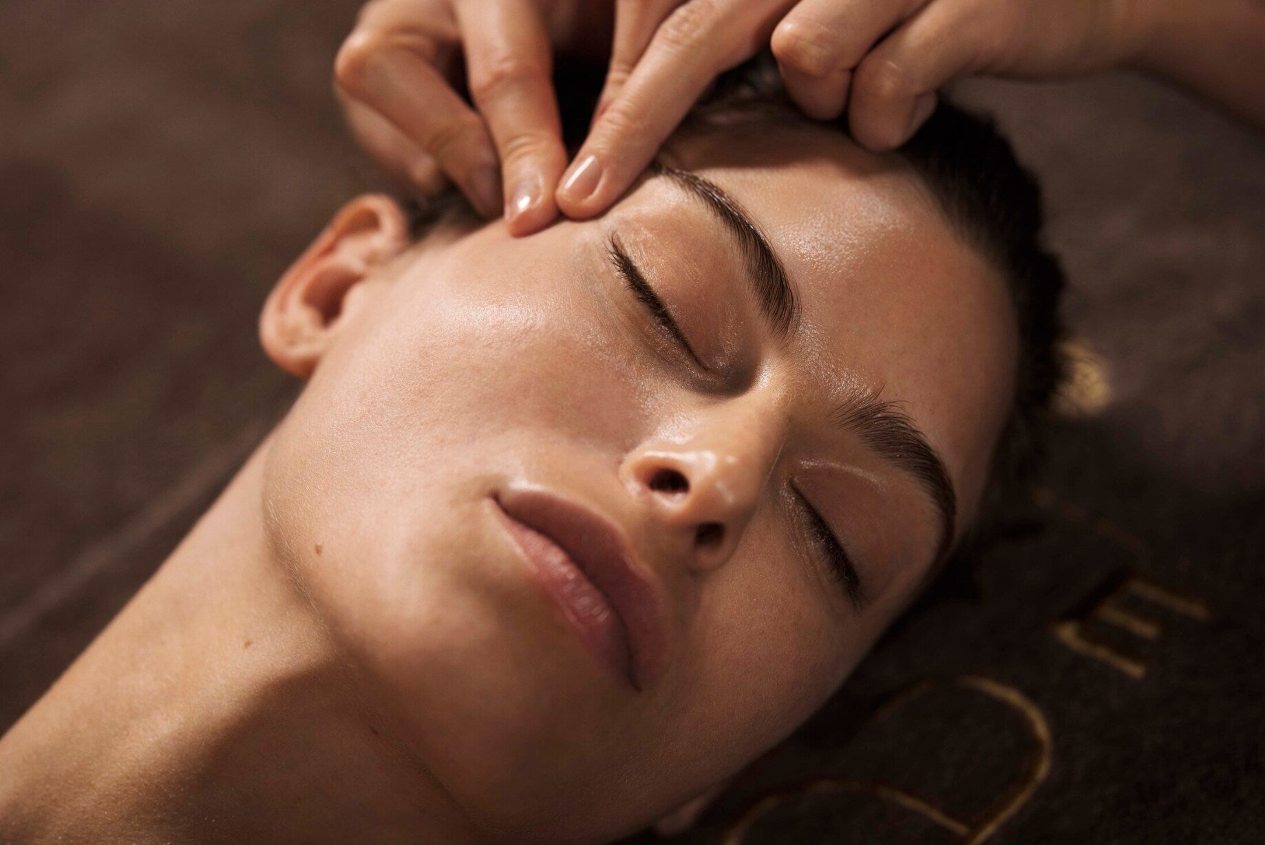 Un visage est montré en gros plan, allongé sur une surface sombre, avec les yeux fermés. Deux doigts exercent une pression précise au niveau de la tempe, juste au-dessus de la joue, dans un geste lent de massage.
La lumière chaude met en valeur la peau lisse et légèrement brillante, ainsi que les contours du nez, des lèvres et du front. L’arrière-plan reste flou, créant une ambiance apaisante et centrée sur le soin du visage.
