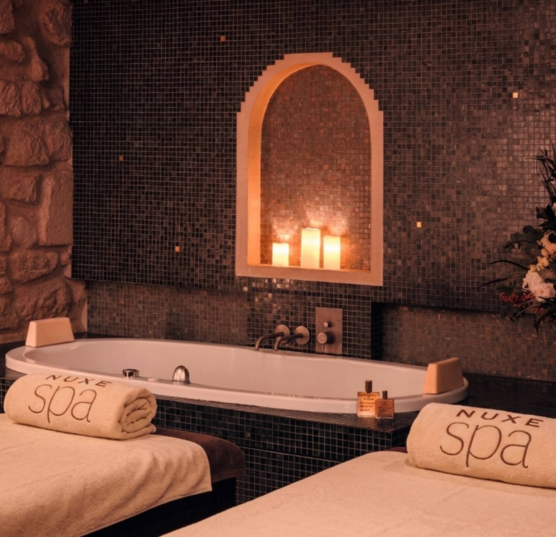 Une baignoire encastrée est placée au centre d’un espace aux murs recouverts de petites mosaïques sombres. Dans une niche en forme d’arche au-dessus, plusieurs bougies allumées diffusent une lumière chaude et tamisée.
Au premier plan, deux tables de massage recouvertes de draps beiges portent des serviettes roulées marquées « NUXE spa ». L’ensemble de la pièce, avec ses pierres apparentes et son éclairage doux, crée une atmosphère intime et relaxante.