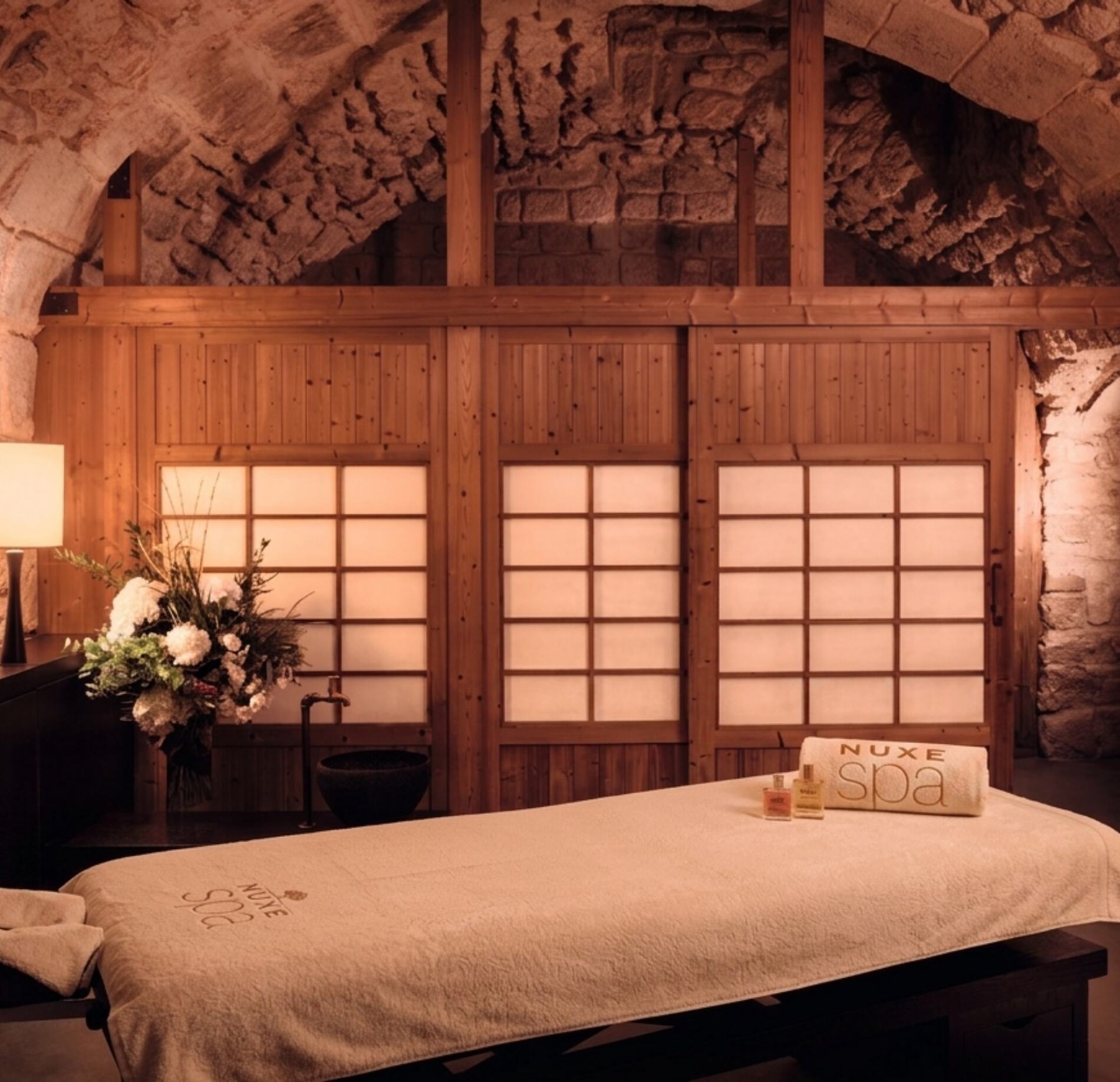 Une table de massage recouverte d’un drap beige occupe le premier plan, avec une serviette roulée marquée « NUXE spa » et deux petits flacons posés dessus.
À l’arrière, des panneaux en bois clair avec des motifs quadrillés filtrent une lumière douce, tandis que des murs en pierre apparente et une voûte renforcent l’aspect chaleureux. L’ensemble crée une ambiance apaisante, mêlant bois, pierre et éclairage tamisé.