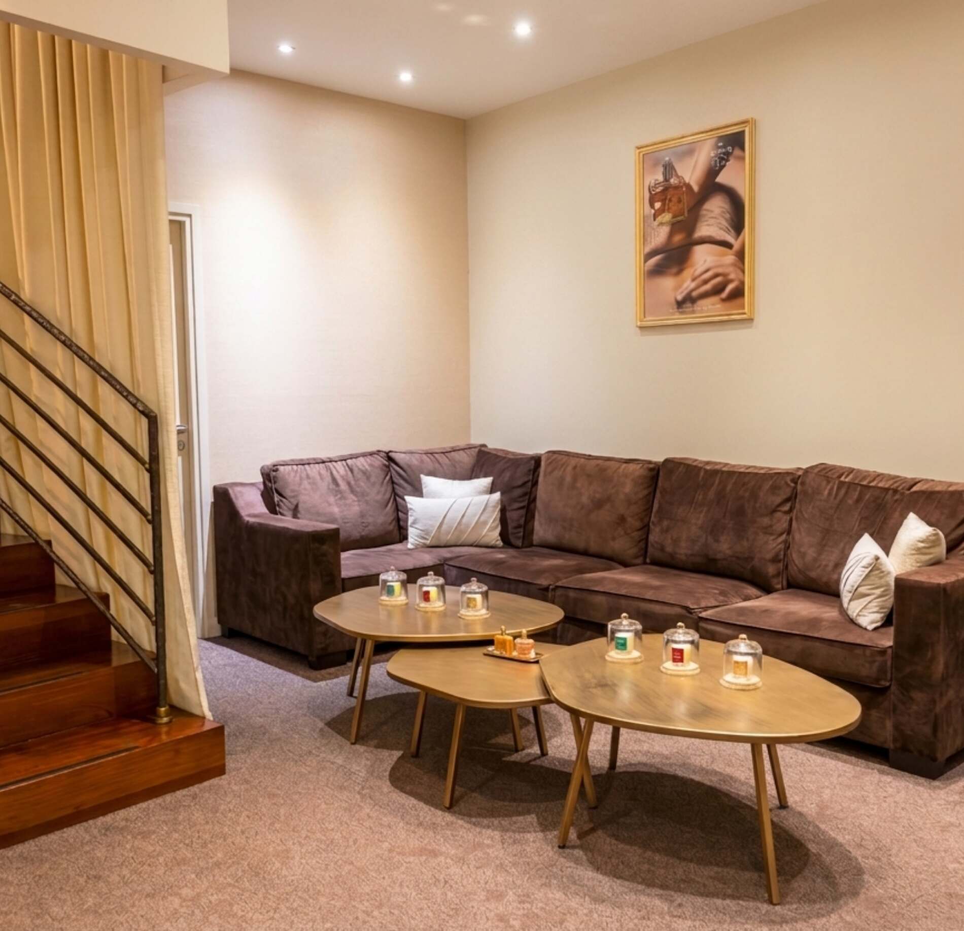 Une pièce de détente est aménagée avec un grand canapé d’angle en tissu marron, entouré de coussins clairs. Devant le canapé, deux tables basses en bois accueillent plusieurs petites bougies parfumées disposées sous des cloches en verre.
Sur le mur, un tableau montre un flacon de parfum tenu au-dessus d’un corps, et un escalier en bois avec rampe métallique est visible sur la gauche. L’ensemble est éclairé par une lumière chaude et douce, créant une ambiance calme et accueillante.
