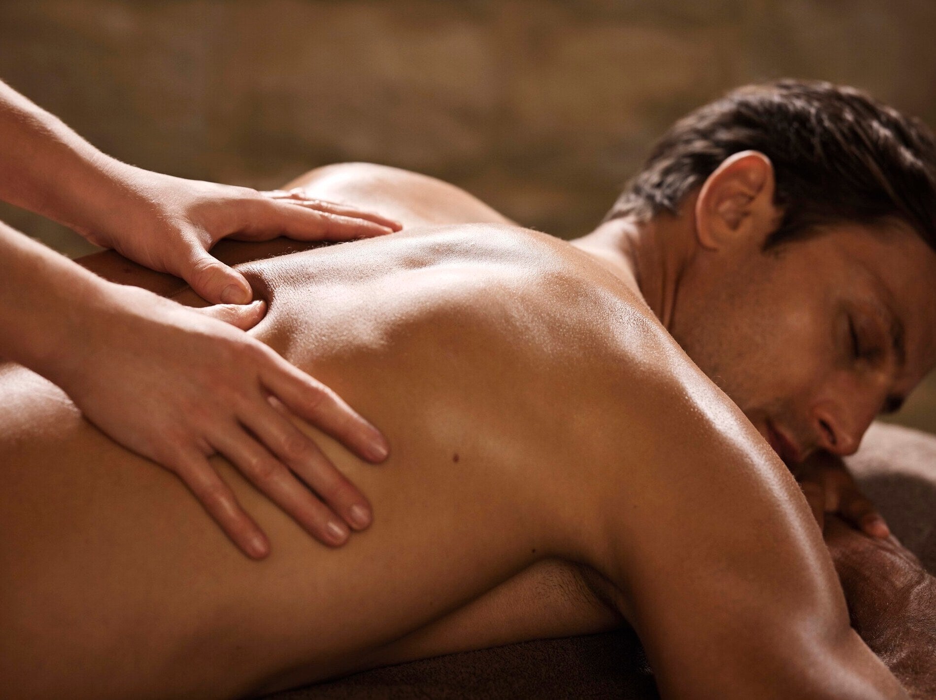 Un homme brun est allongé de profil sur le ventre, les yeux clos et le visage détendu. Deux mains massent doucement le côté de son dos nu, dont la peau est légèrement brillante, sous un éclairage chaleureux et enveloppant propre à un spa.