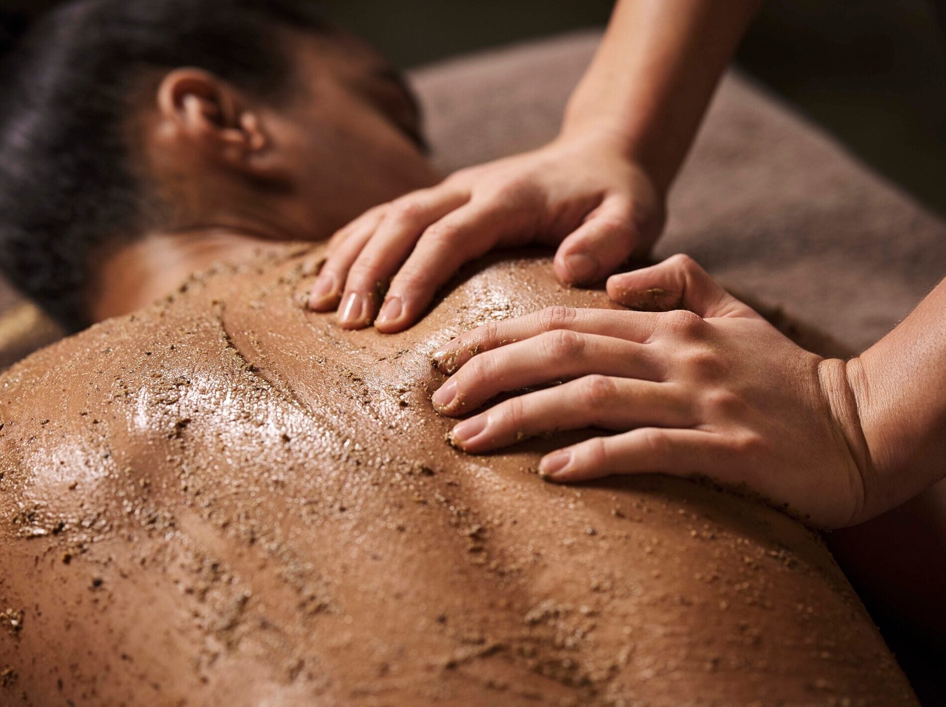 Une personne est allongée sur le ventre pendant qu'on applique sur son dos un gommage texturé aux grains fins et marron. Deux mains étalent ce soin exfoliant sur la peau humide, créant une sensation de soin corporel dans une ambiance tamisée.