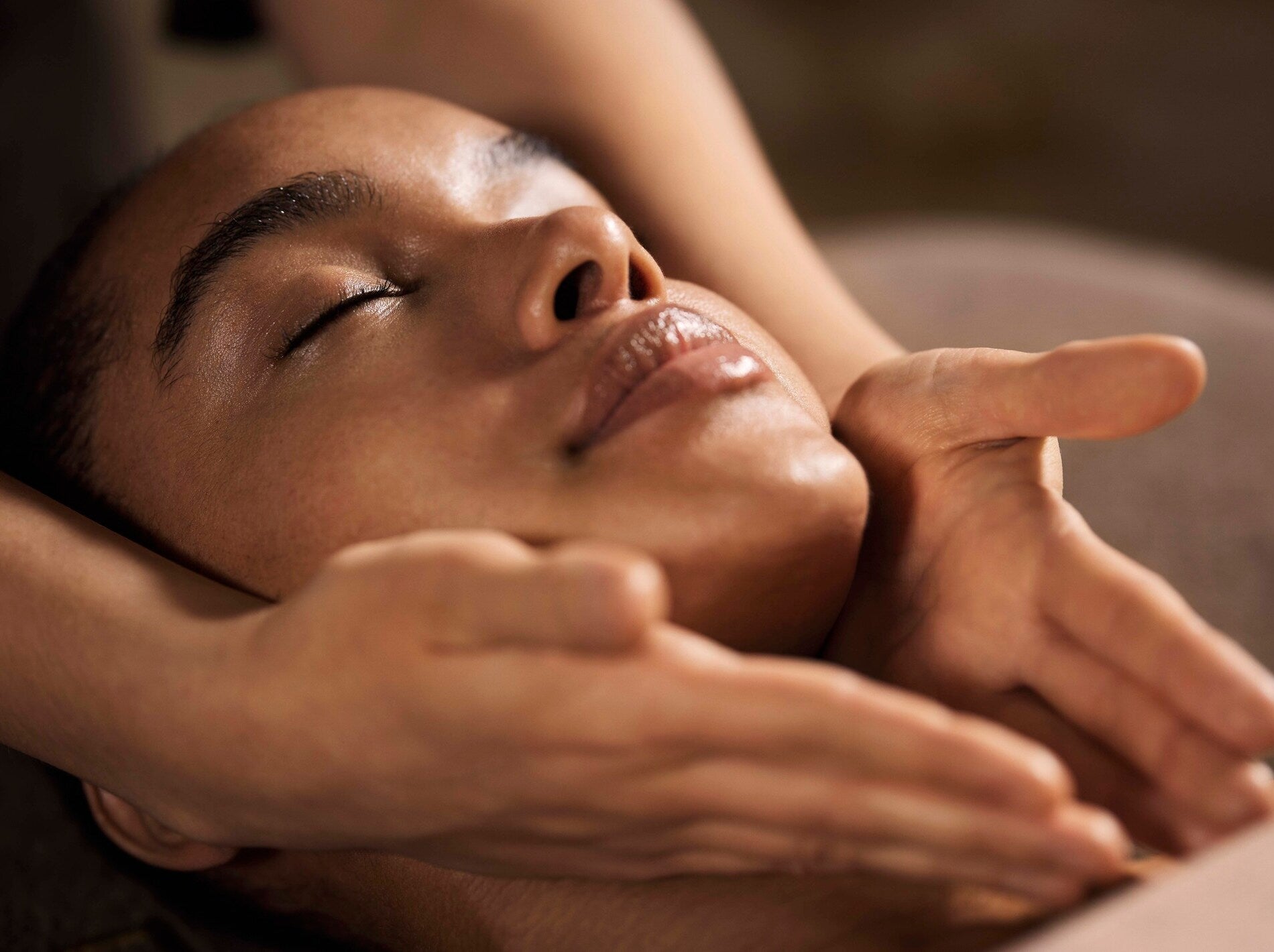 Un visage est montré en gros plan, allongé avec les yeux fermés, la tête légèrement inclinée vers l’arrière. Deux mains entourent délicatement le bas du visage, en soutenant le menton dans un geste lent et précis de massage.
La lumière chaude met en valeur la peau lisse et brillante ainsi que les contours du nez et des lèvres. L’arrière-plan est flou et doux, renforçant l’ambiance apaisante et soignée d’un rituel de spa.