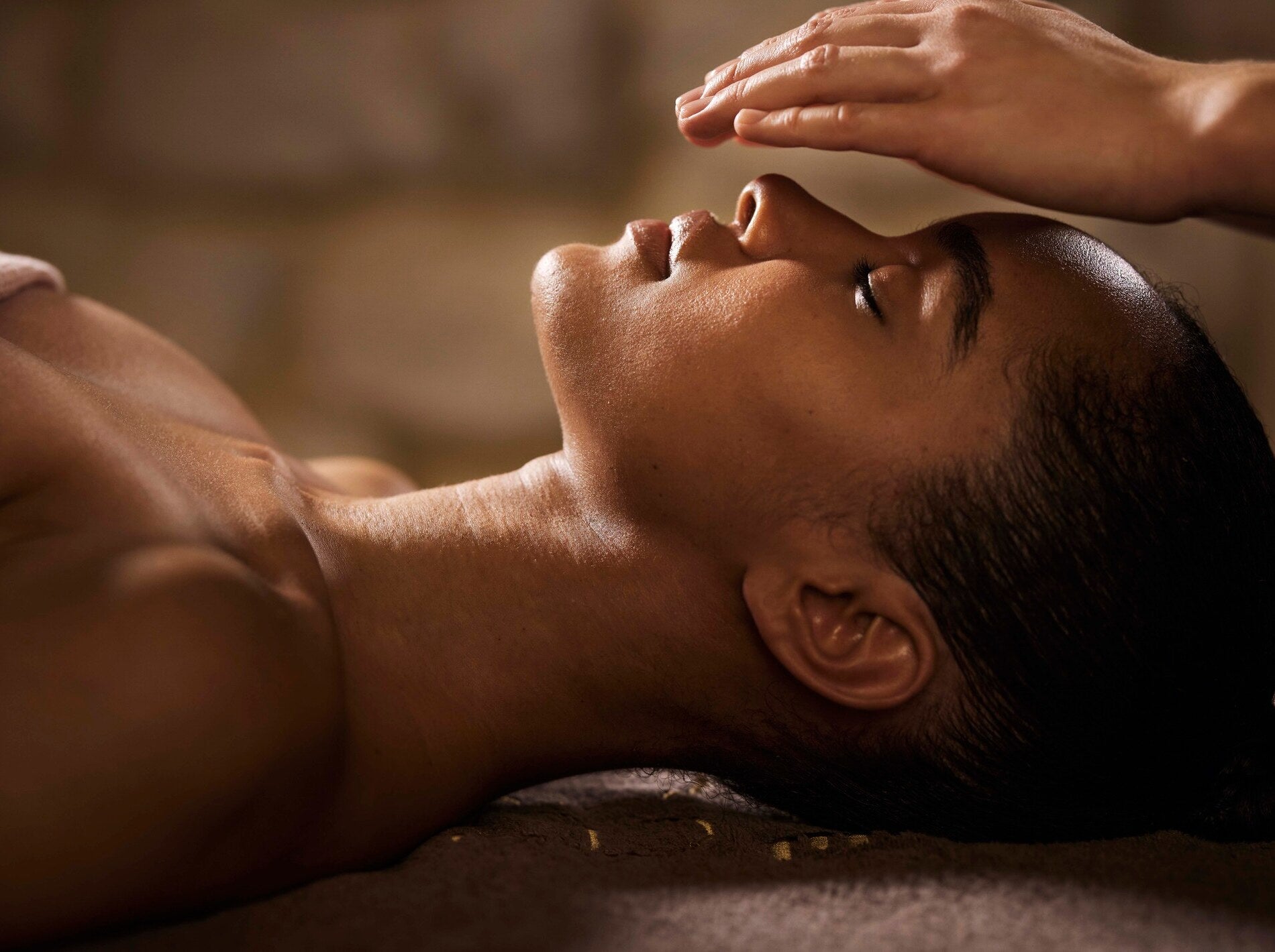 Une personne est allongée de profil sur une surface sombre, le visage tourné vers le haut avec les yeux fermés. Une main est posée doucement sur le front, dans un geste lent et contrôlé de massage.
La lumière chaude souligne les contours du visage, du cou et de l’épaule, avec une peau légèrement brillante. L’arrière-plan est flou, créant une atmosphère calme et apaisante de soin en spa.