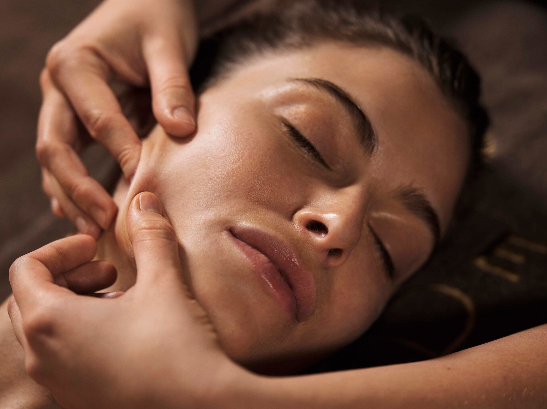 Un visage est visible en gros plan, allongé sur une surface sombre, avec les yeux fermés. Deux mains entourent le bas du visage et exercent une pression douce le long de la mâchoire, dans un mouvement précis de massage.
La lumière chaude fait ressortir la texture lisse et légèrement brillante de la peau. L’arrière-plan reste flou, renforçant l’atmosphère calme et apaisante d’un soin en spa.
