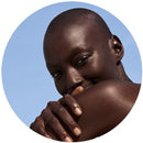 visage d'une femme, légèrement cachée par sa propre épaule. Elle est chauve, les yeux marrons et regarde l'objectif 