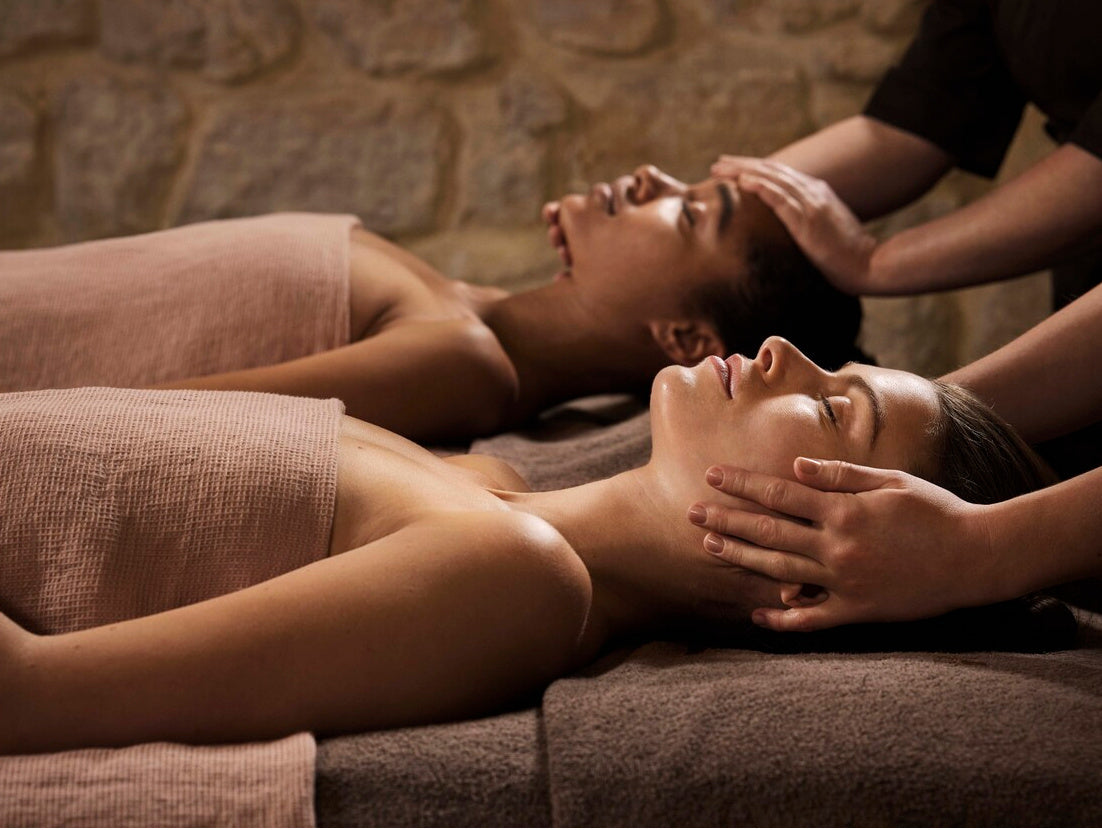 Deux femmes profitent d'un massage crânien et facial en duo, allongées sur le dos, le corps drapé de serviettes roses. Au premier plan, des mains massent délicatement le visage d'une femme rousse aux yeux fermés, tandis qu'à l'arrière-plan, une autre femme reçoit le même soin dans une pièce tamisée en pierres apparentes.