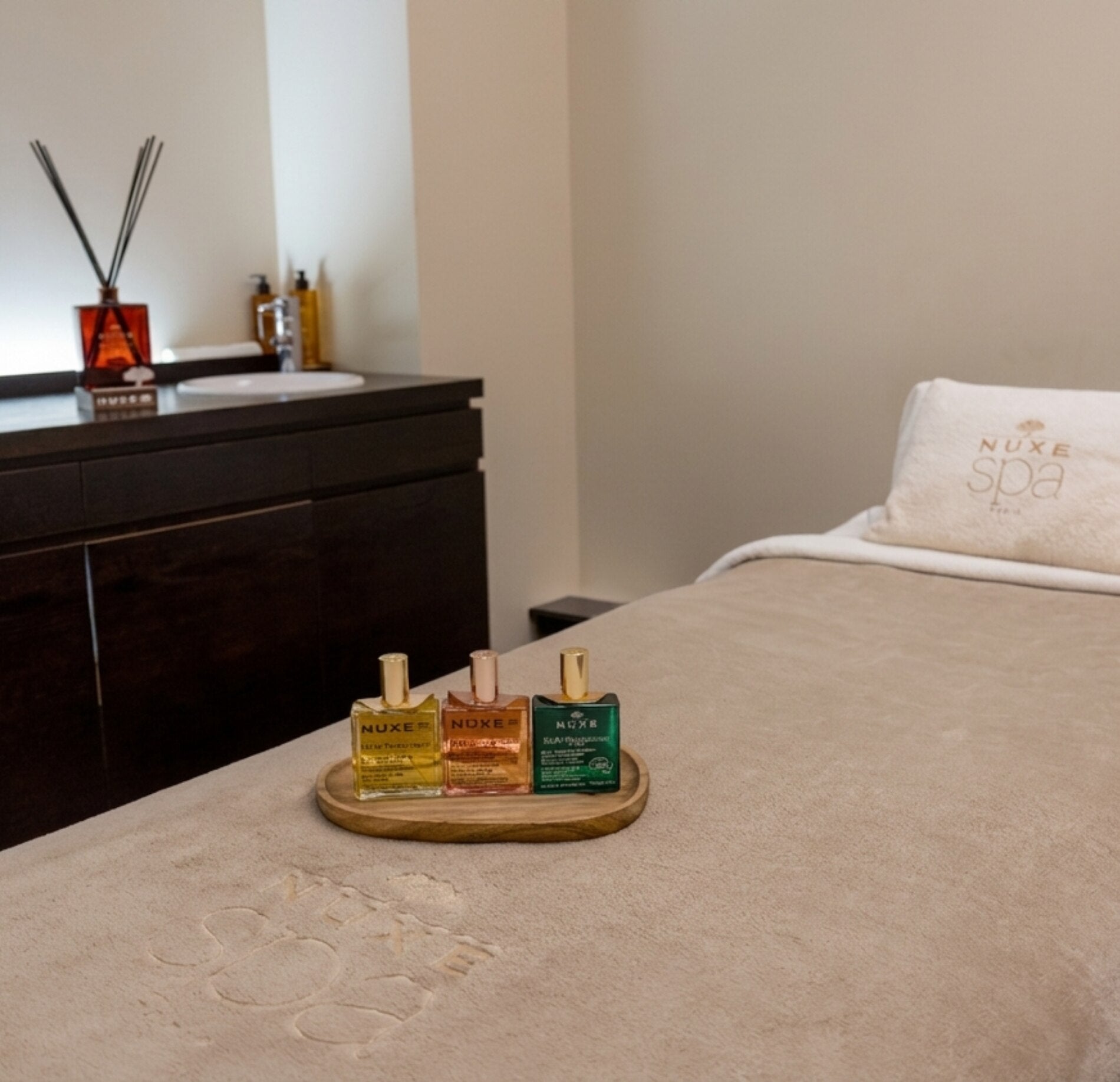 Une table de massage recouverte d’un drap beige occupe le centre de la pièce, avec trois petits flacon Nuxe plus précisément trois petites Huile prodigieuse. Sur l’oreiller, une serviette blanche porte l’inscription « NUXE spa ».
À l’arrière-plan, un meuble sombre accueille un diffuseur avec des tiges et quelques bouteilles, près d’un lavabo. L’ensemble est éclairé par une lumière douce, dans une pièce épurée aux tons clairs et apaisants.