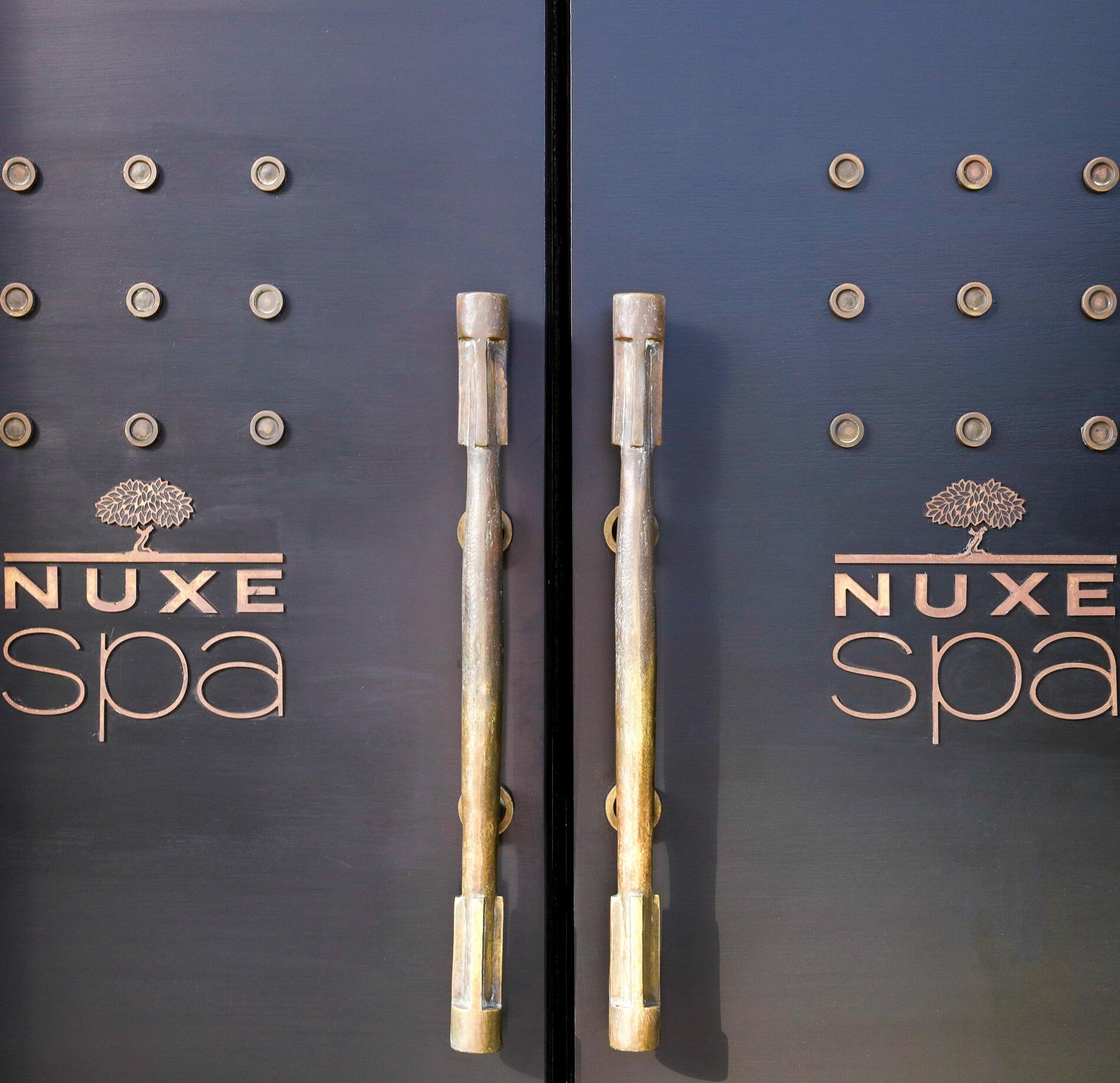 Deux grandes portes sombres occupent toute l’image, avec des poignées verticales en métal au centre. Chaque porte est décorée de petits reliefs circulaires disposés en grille, donnant un aspect texturé et élégant.
Le logo « NUXE spa » est inscrit en lettres dorées sur les deux battants, avec un arbre stylisé au-dessus du mot. L’ensemble est éclairé doucement, mettant en valeur le contraste entre le noir mat des portes et les reflets dorés des inscriptions et des poignées.