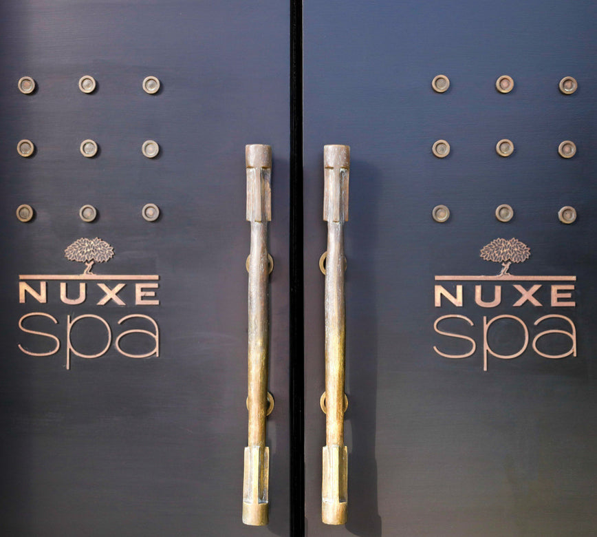 Deux grandes portes sombres occupent toute l’image, avec des poignées verticales en métal au centre. Chaque porte est décorée de petits reliefs circulaires disposés en grille, donnant un aspect texturé et élégant.
Le logo « NUXE spa » est inscrit en lettres dorées sur les deux battants, avec un arbre stylisé au-dessus du mot. L’ensemble est éclairé doucement, mettant en valeur le contraste entre le noir mat des portes et les reflets dorés des inscriptions et des poignées.