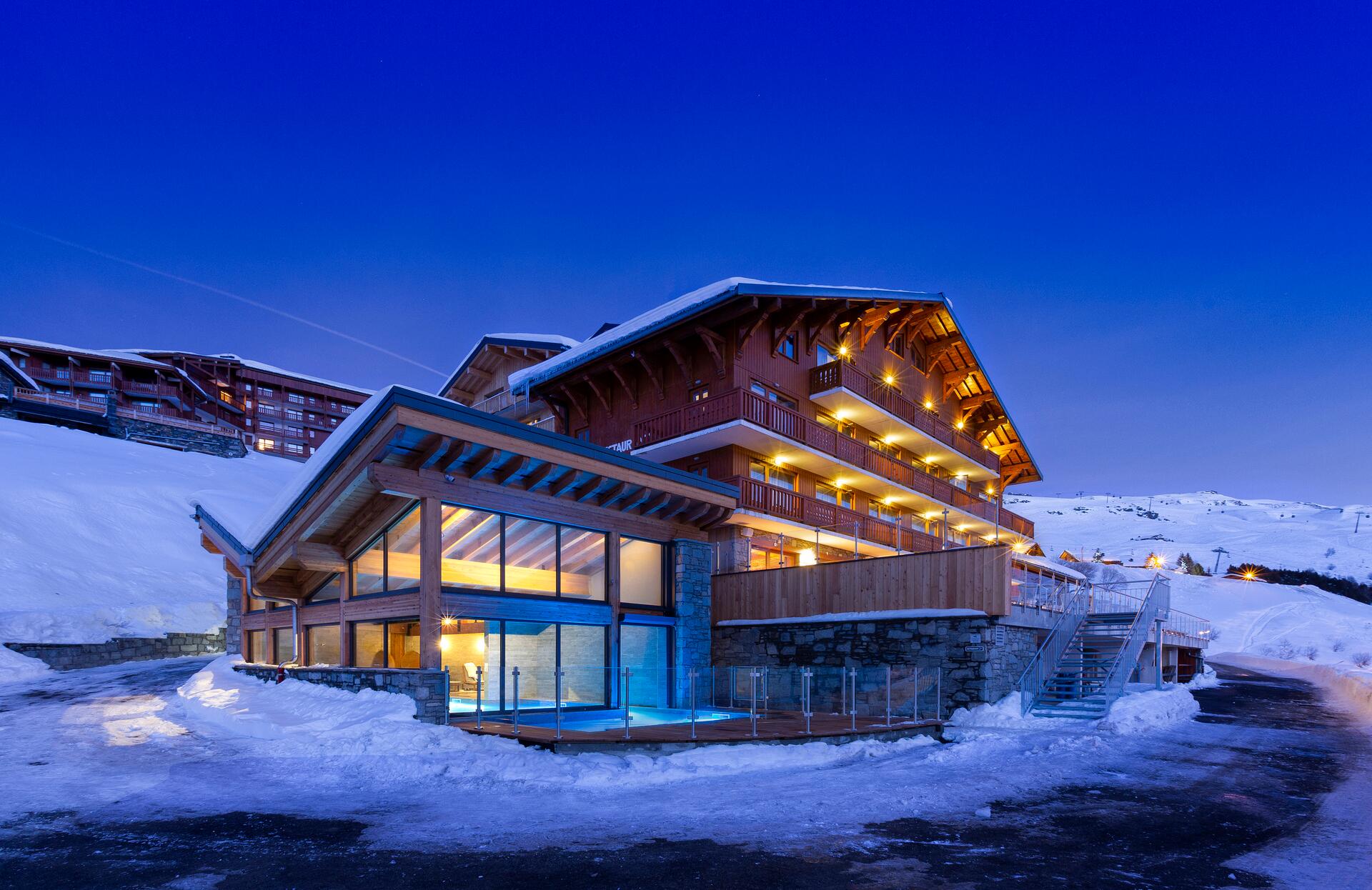 Un chalet alpin majestueux se dresse au cœur d’un paysage enneigé, avec des façades en bois chaleureux illuminées par des lumières dorées contrastant avec le ciel bleu profond du soir.
Au pied du bâtiment, un espace vitré révèle une piscine intérieure baignée de lumière douce, offrant une expérience cosy et luxueuse dans un décor naturel spectaculaire.