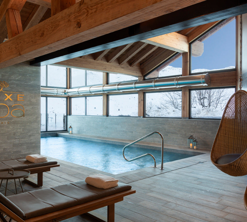 Une piscine intérieure du spa de l'hotel l'Ours Blanc s’étend dans une pièce lumineuse avec une charpente en bois apparente et de grandes baies vitrées.
Des transats avec serviettes sont disposés au bord du bassin, tandis que la vue donne sur un paysage enneigé, créant une ambiance calme et naturelle.