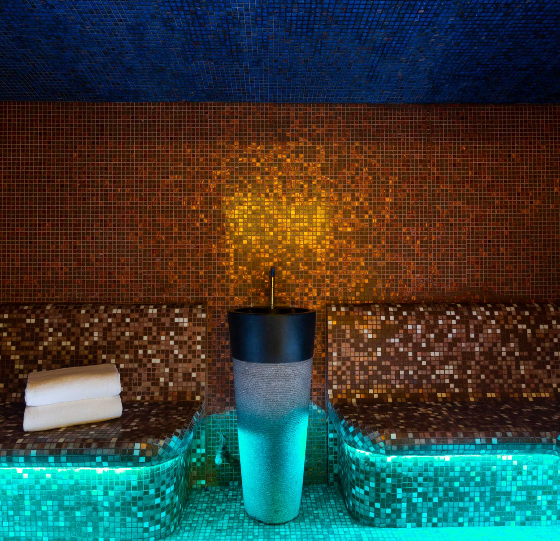 Un hammam du spa de l'hotel l'Ours Blanc est recouvert de mosaïques colorées, avec des bancs arrondis éclairés par une lumière bleutée en dessous.
Au centre, une fontaine moderne diffuse de l’eau, entourée de serviettes roulées, créant une ambiance chaleureuse et immersive mêlant tons dorés et bleus.